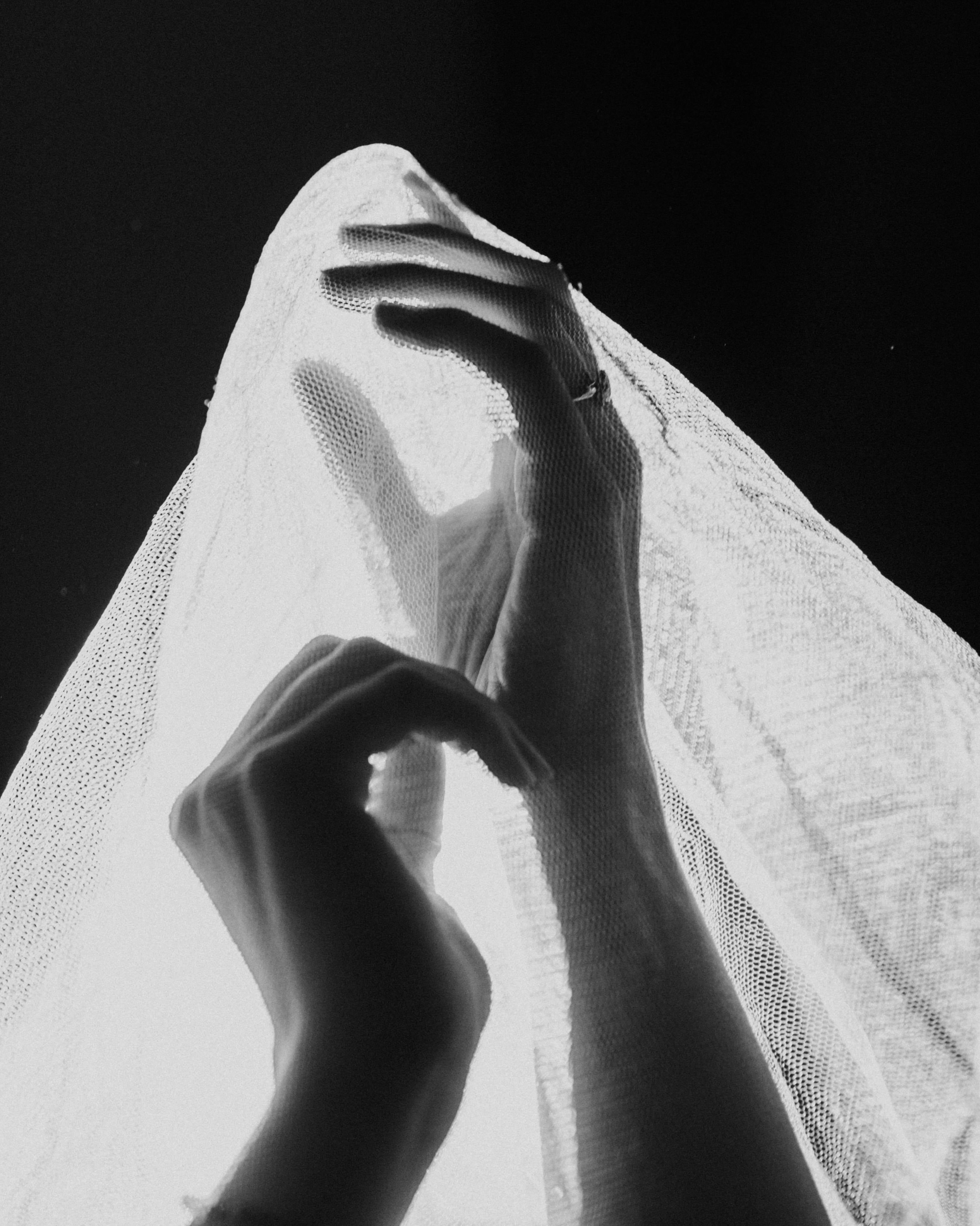A black and white photo of two hands reaching upwards wearing rings and are covered in a transparent white veil.