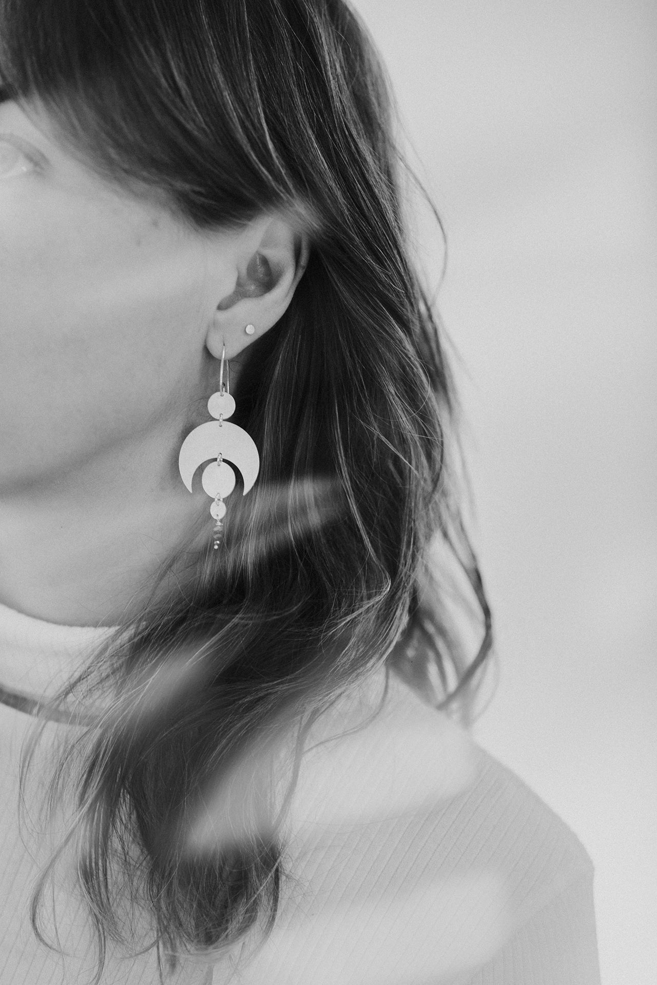 A black and white image of the side profile of a person with long hair. They are wearing the Celestial Earrings by Emily Eliza Arlotte Handcrafted Fine Jewellery. The earrings are large and consist of sterling silver moon and circle shapes.