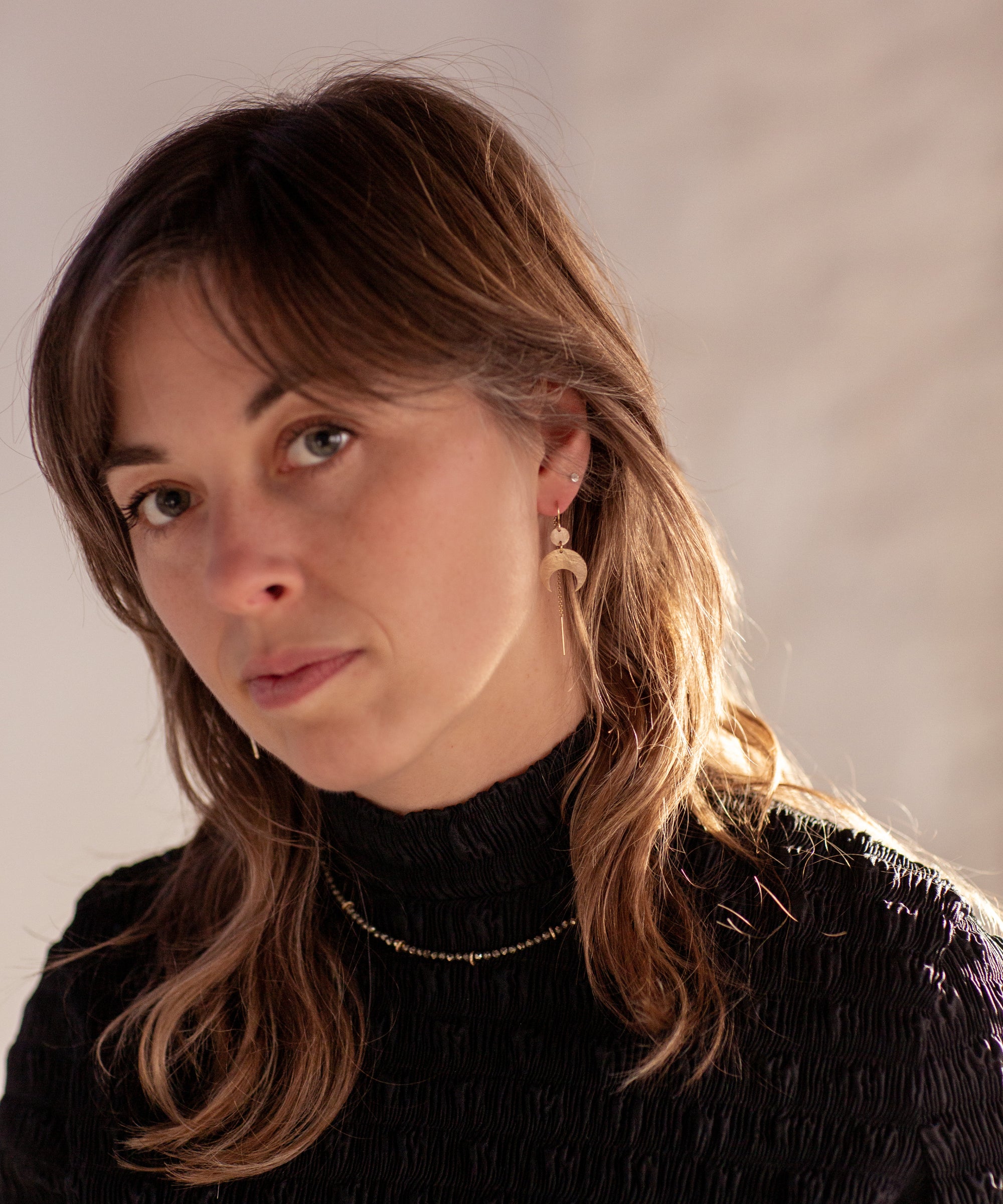 Woman with long brown hair wearing a black turtleneck, Gold Lunar Crescent Moon Thread Earrings, and a Pyrite Choker Necklace against a neutral background