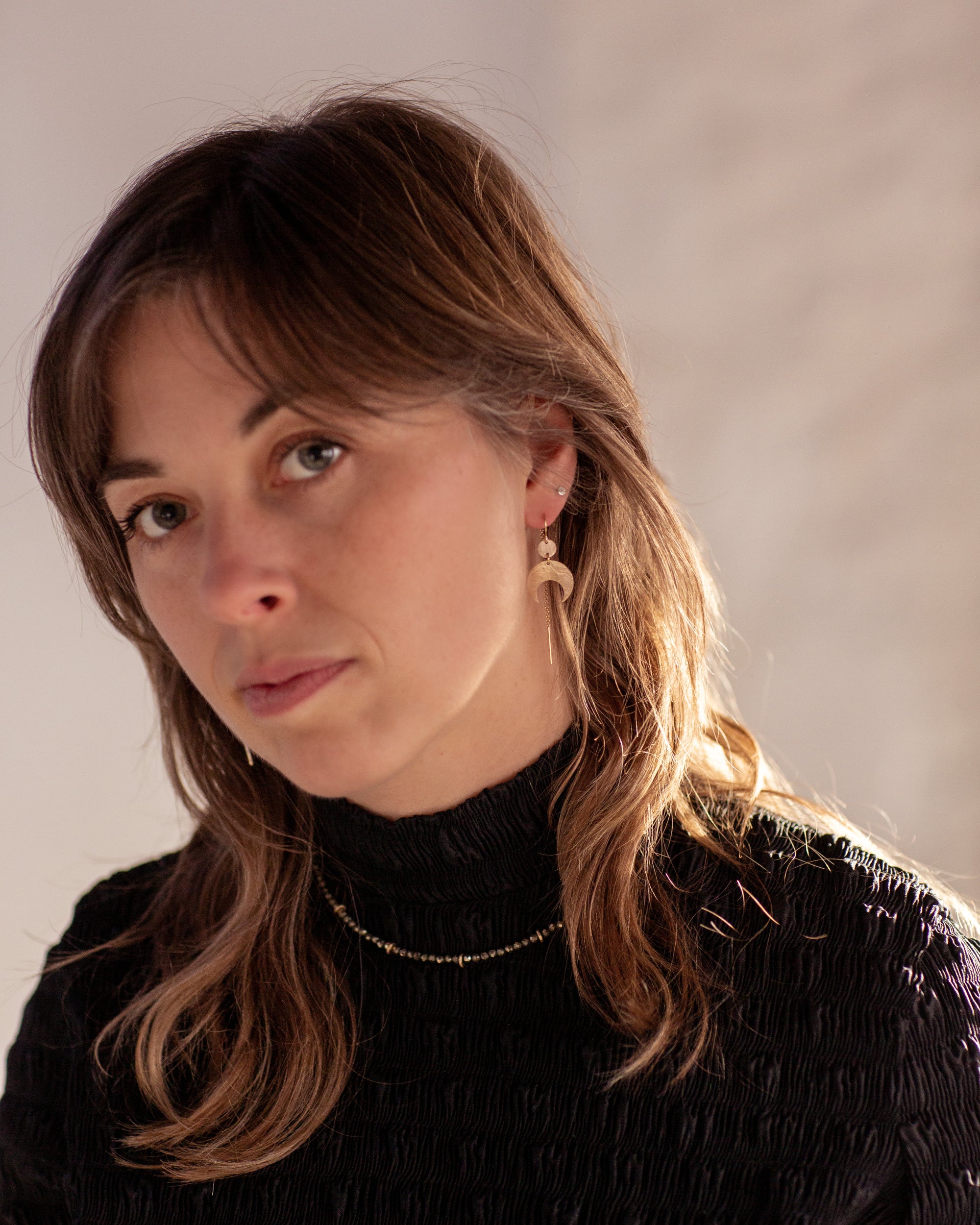 Woman with long brown hair wearing a black turtleneck, Gold Lunar Crescent Moon Thread Earrings, and a Pyrite Choker Necklace against a neutral background
