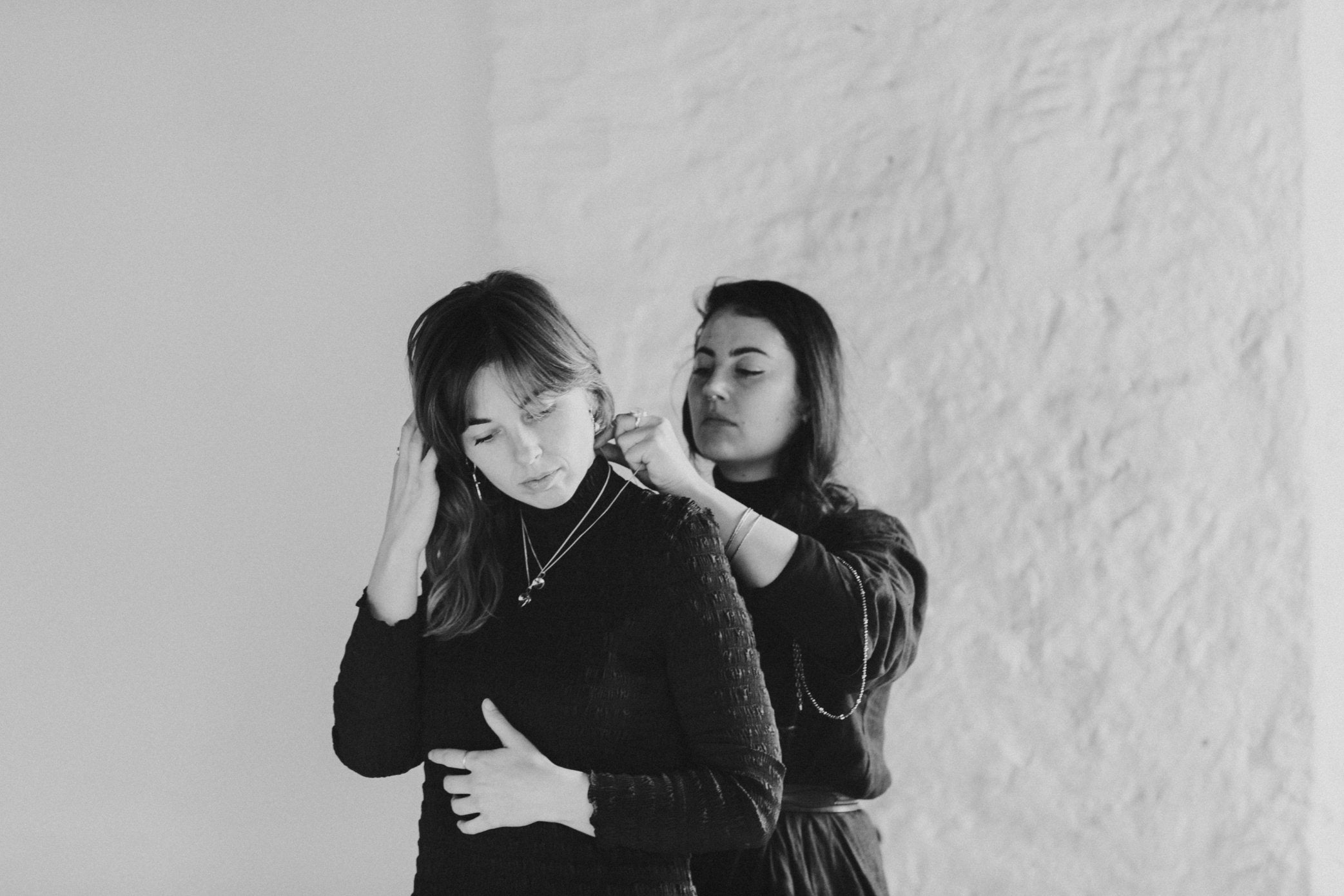 A black and white shot of a behind the scenes moment at a jewellery editorial photoshoot. Emily Eliza Arlotte helps model Eden try on necklaces.