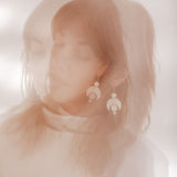 A double exposure photograph of a woman wearing the Celestial earrings. The photo is in soft pink and browns. The earrings featuring a crescent moon and circles representing planetary alignment.