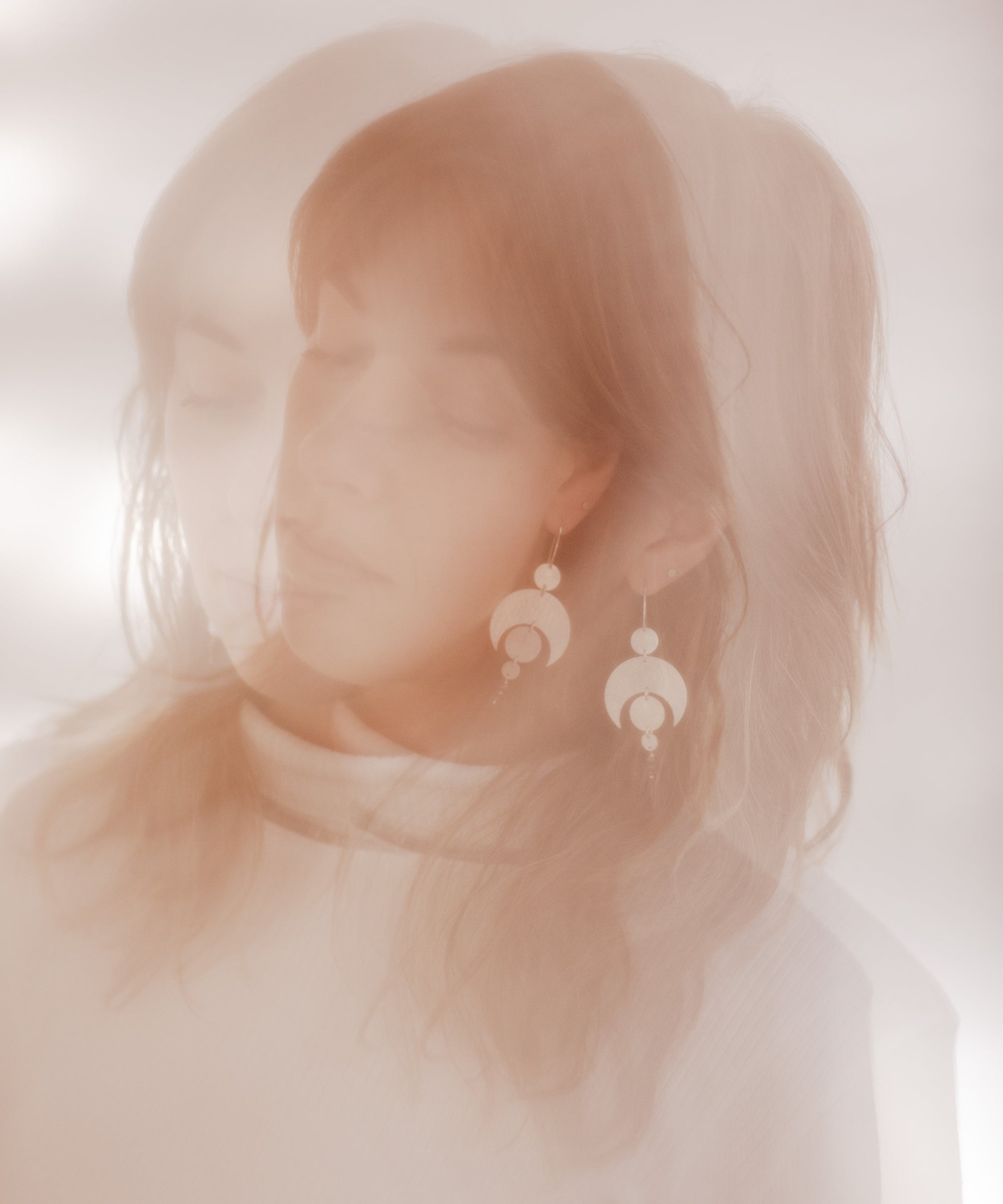 A double exposure photograph of a woman wearing the Celestial earrings. The photo is in soft pink and browns. The earrings featuring a crescent moon and circles representing planetary alignment.