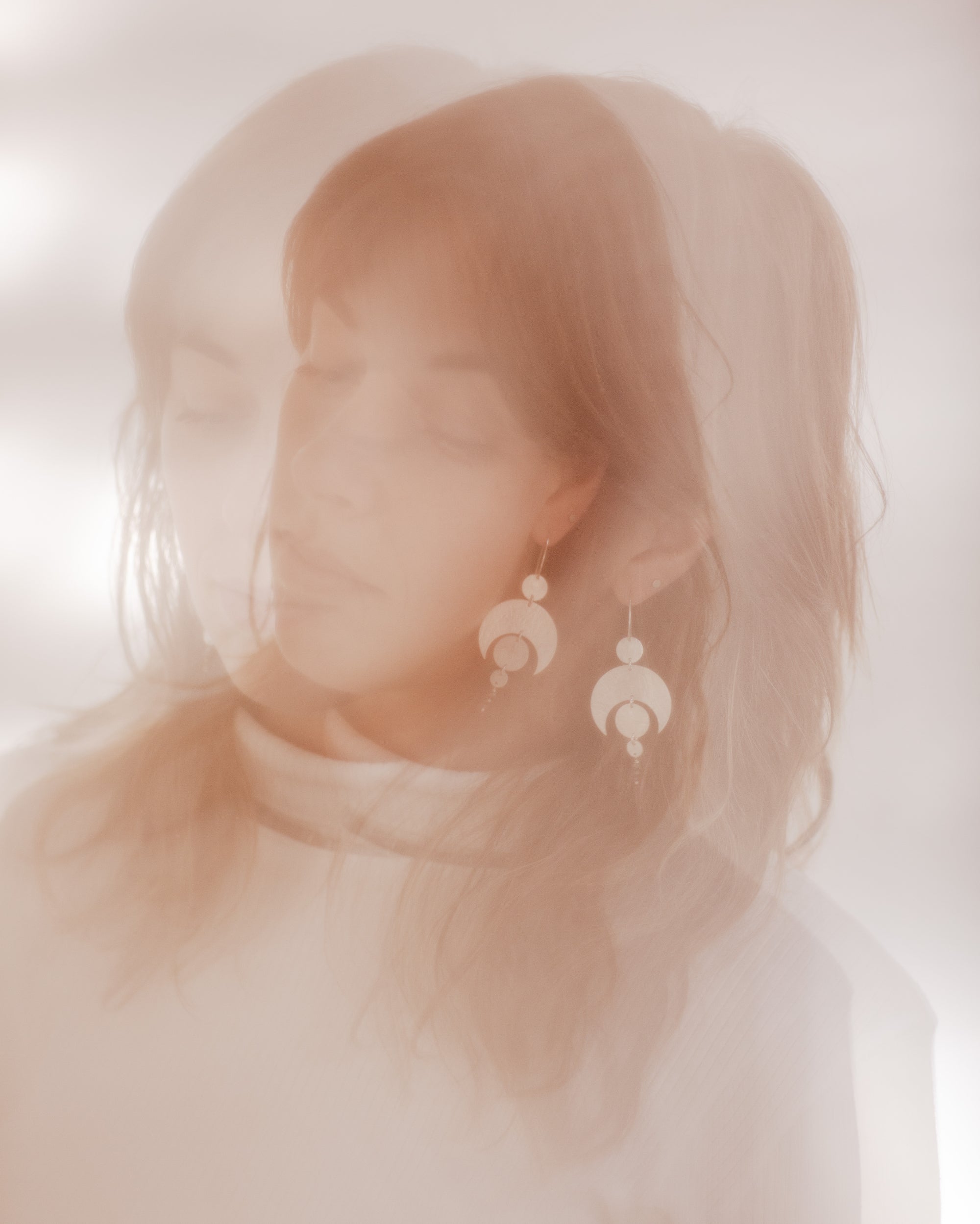 A double exposure photograph of a woman wearing the Celestial earrings. The photo is in soft pink and browns. The earrings featuring a crescent moon and circles representing planetary alignment.
