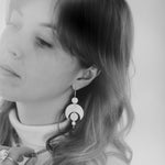 Black and white photo of a person wearing the Celestial Earrings, which feature a moon crescent and circles representing planetary alignment. They also wear a labradorite choker necklace and large quartz ring.