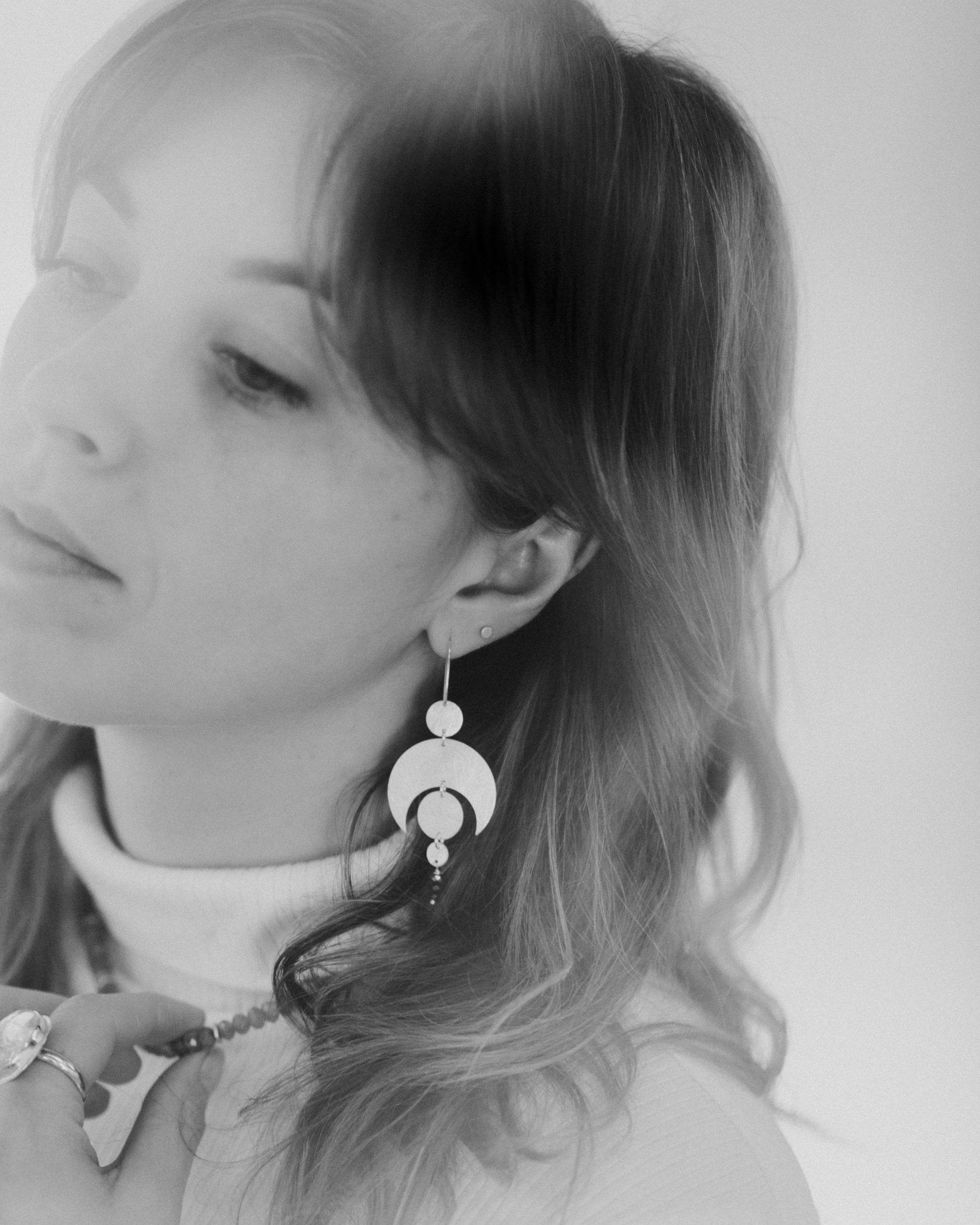 Black and white photo of a person wearing the Celestial Earrings, which feature a moon crescent and circles representing planetary alignment. They also wear a labradorite choker necklace and large quartz ring.