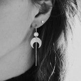 Close-up of a person wearing the Lunar Thread Earrings. These sterling silver earrings feature a textured disc and crescent moon hanging from a hook and fine chain fitting.