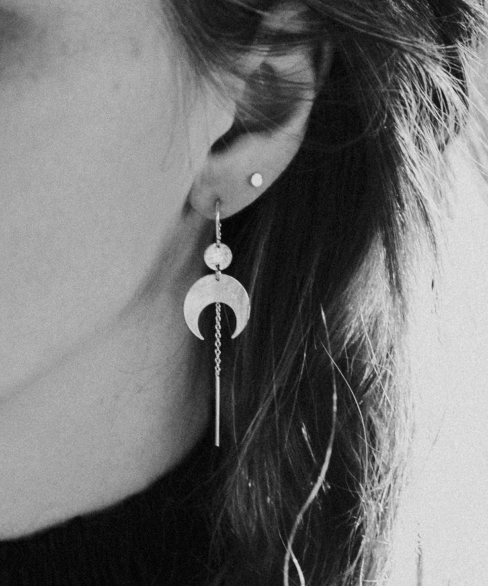 Close-up of a person wearing the Lunar Thread Earrings. These sterling silver earrings feature a textured disc and crescent moon hanging from a hook and fine chain fitting.