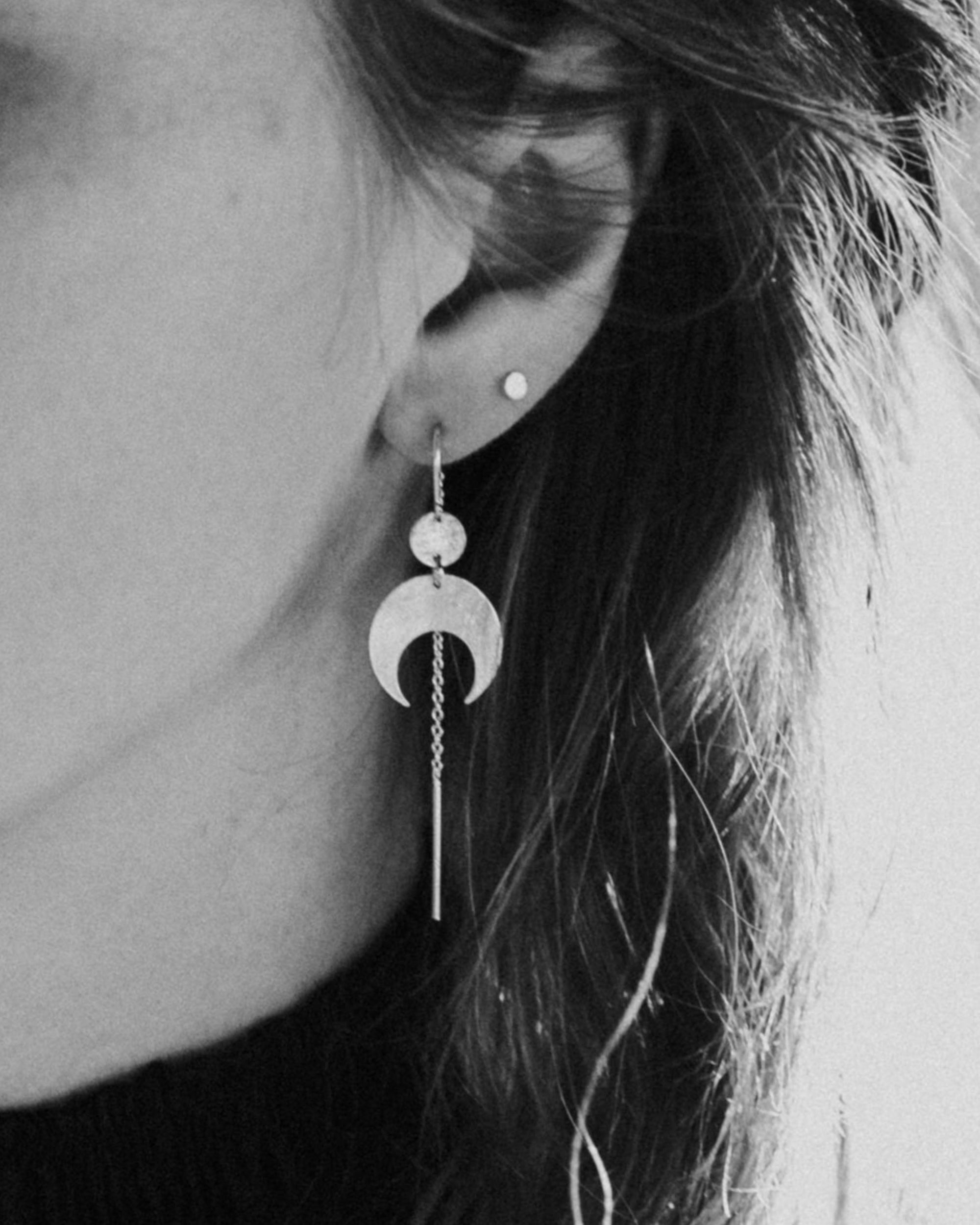 Close-up of a person wearing the Lunar Thread Earrings. These sterling silver earrings feature a textured disc and crescent moon hanging from a hook and fine chain fitting.