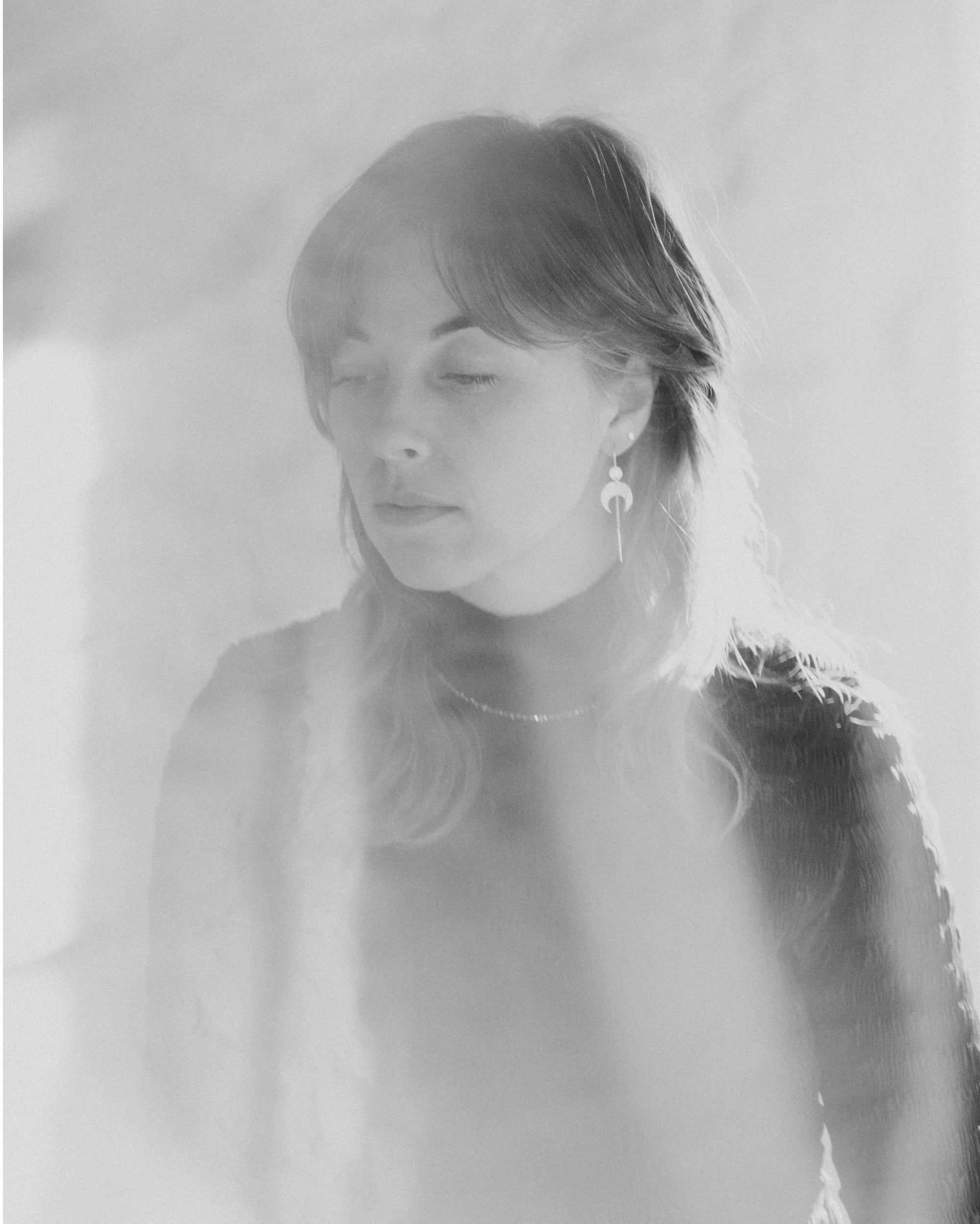 Black and white portrait of a person with shoulder-length hair against a light background. They are wearing the Lunar Thread Earrings. These sterling silver earrings feature a textured disc and crescent moon hanging from a hook and fine chain fitting.