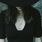 Laska wearing a wide brim black hat and black top and the Luna Crescens Necklace layered with a Butterfly Eclipse Pendant, standing against a blurred background.