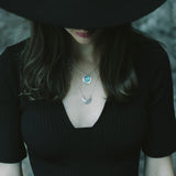 Laska wearing a wide brim black hat and black top and the Luna Crescens Necklace layered with a Butterfly Eclipse Pendant, standing against a blurred background.
