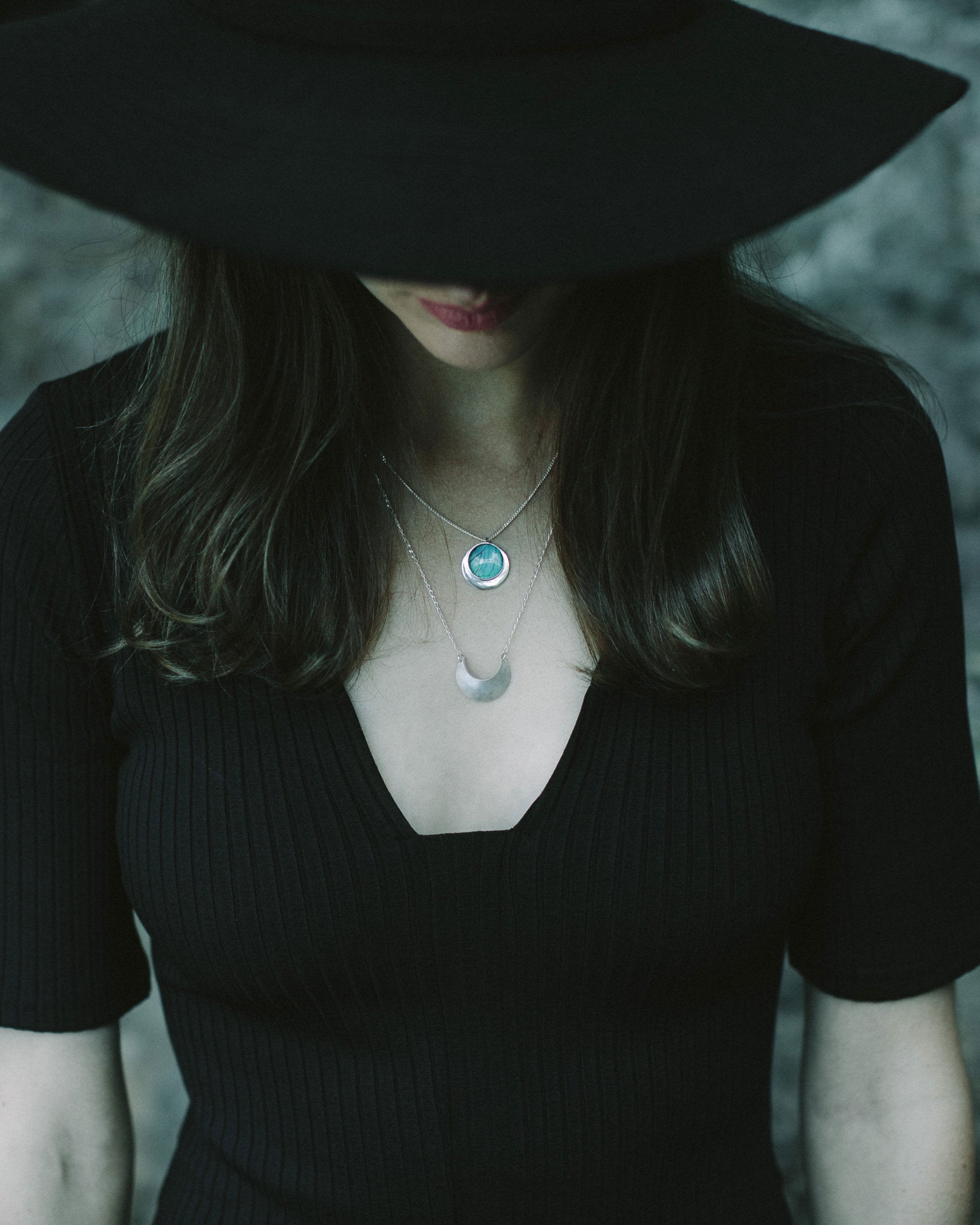 Laska wearing a wide brim black hat and black top and the Luna Crescens Necklace layered with a Butterfly Eclipse Pendant, standing against a blurred background.