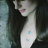 Laska wearing a wide brim black hat and black top and the Luna Crescens Necklace layered with a Butterfly Eclipse Pendant, standing against a rockwall background.