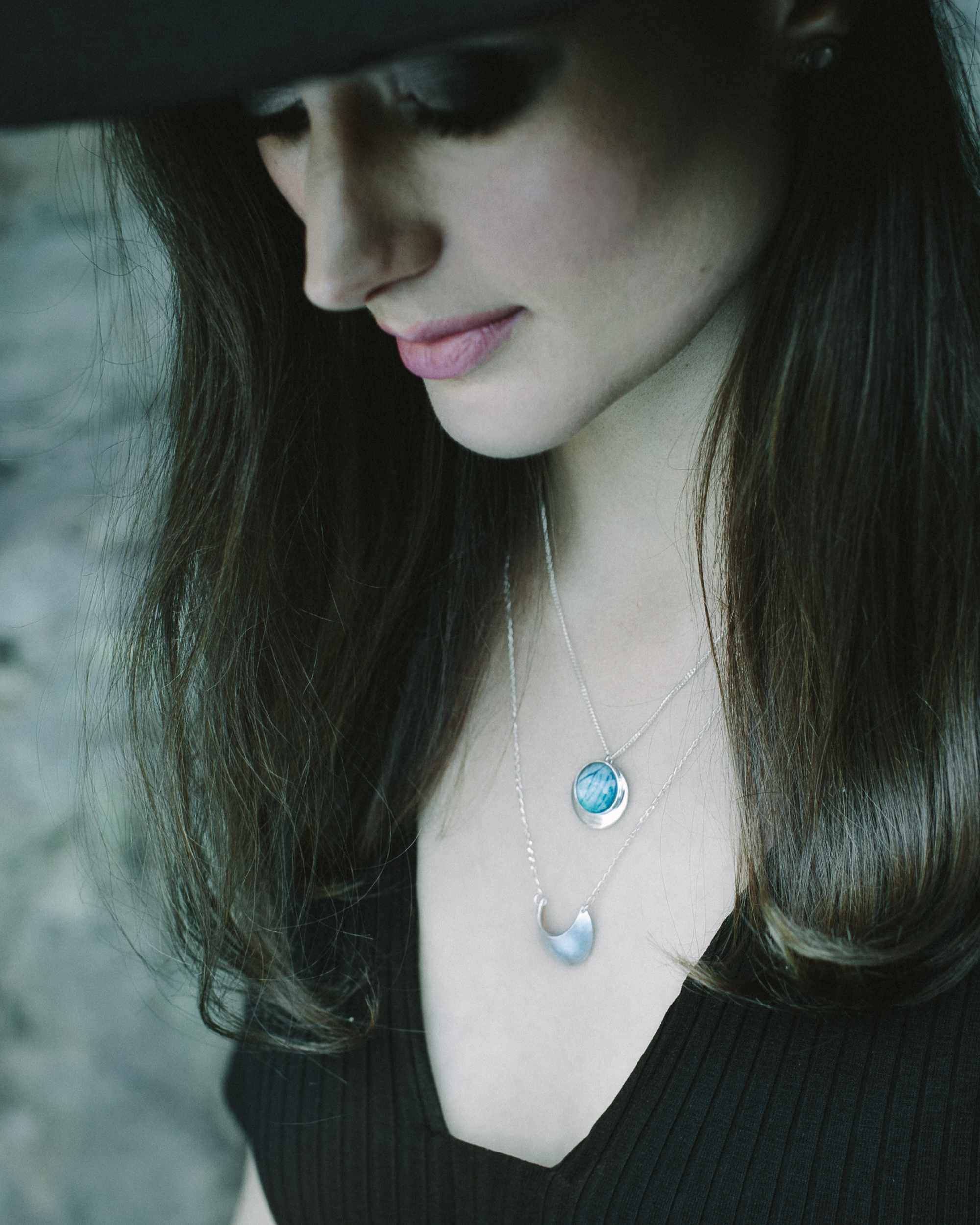 Laska wearing a wide brim black hat and black top and the Luna Crescens Necklace layered with a Butterfly Eclipse Pendant, standing against a rockwall background.