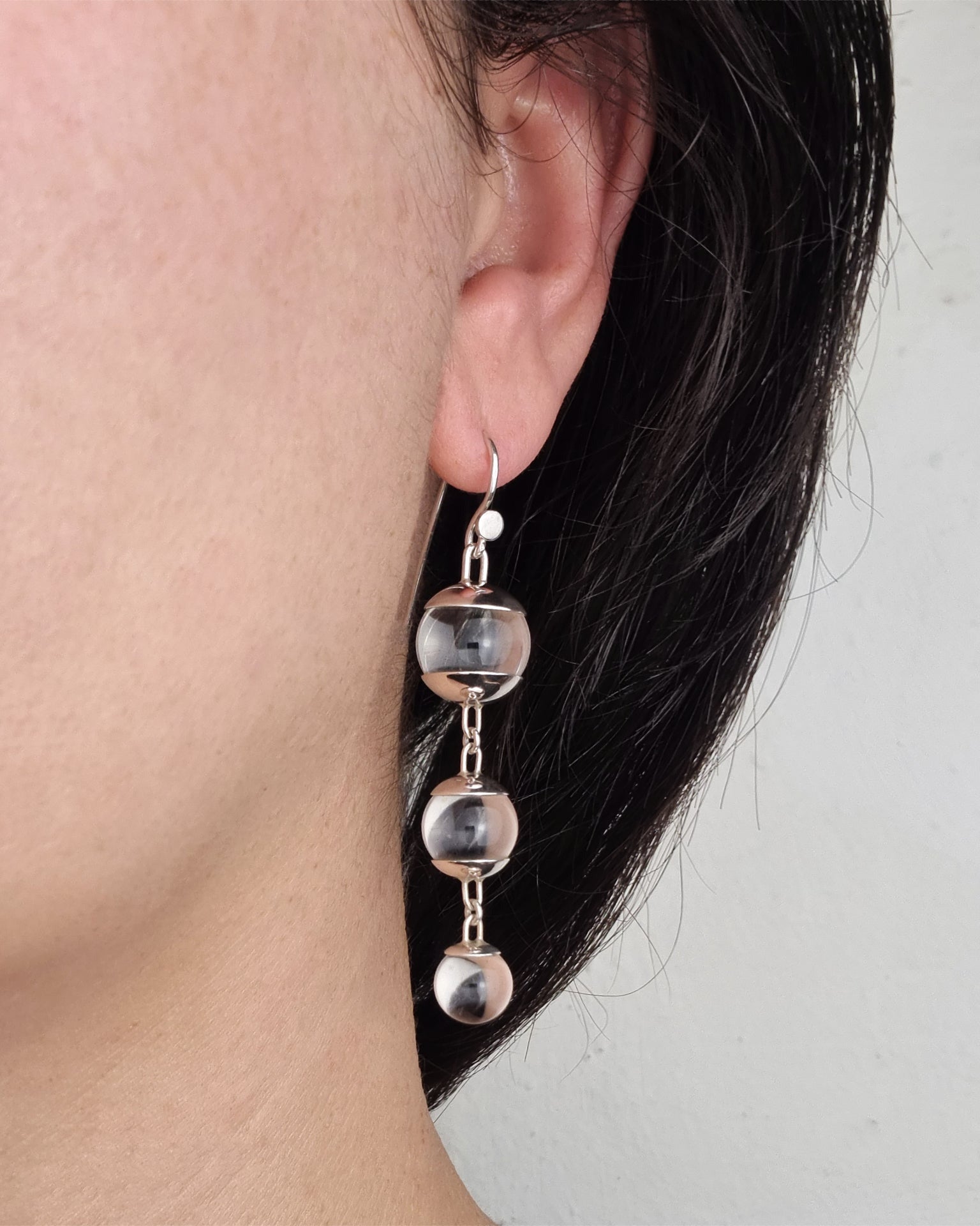 A pair of sterling silver drop earrings featuring three quartz spheres attached to small silver hooks, worn by a person with dark hair.