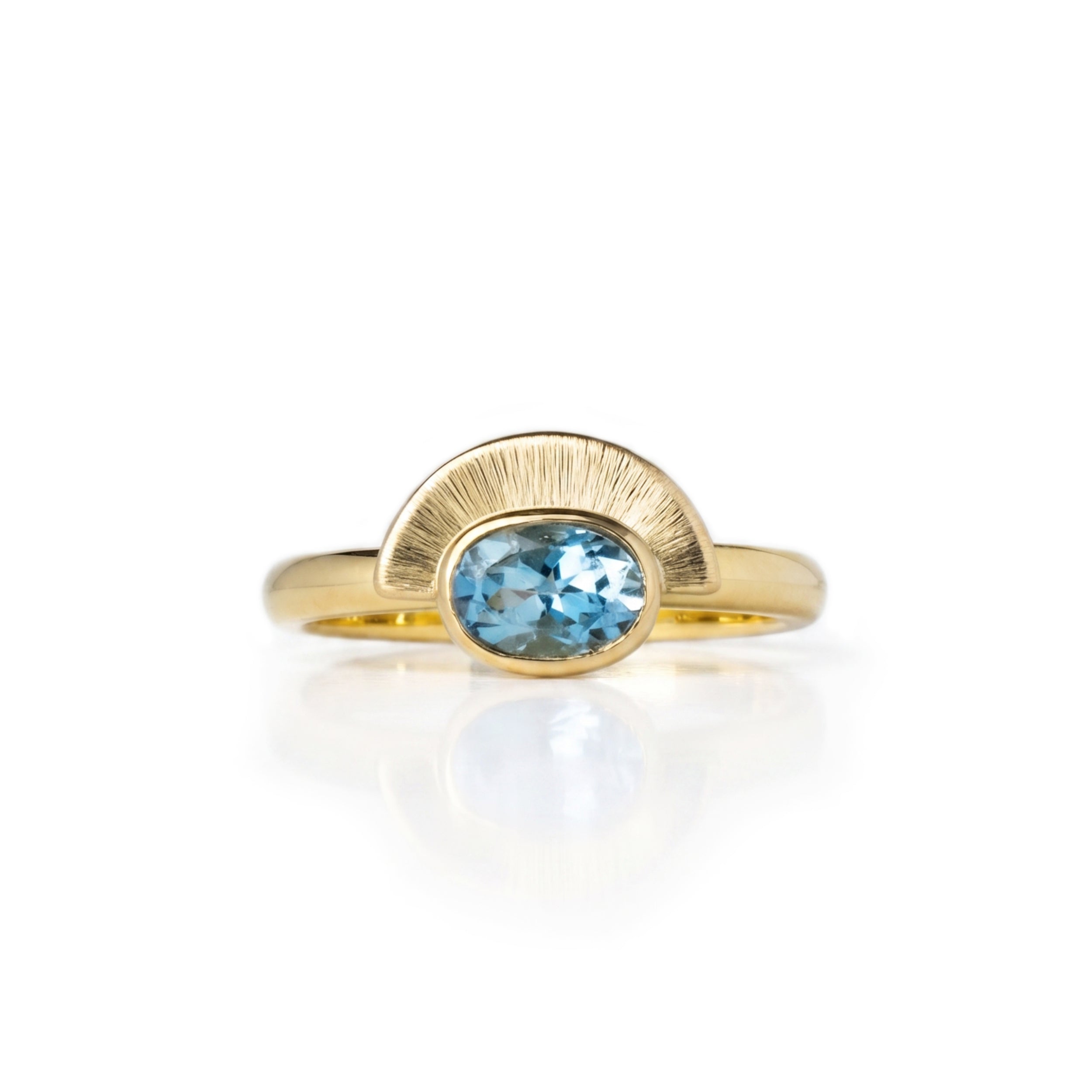 A gold engagement ring featuring an engraved semi-circular arch setting with an oval east west setting Aquamarine, displayed on a clean white background with a soft reflection below. Handmade by Emily Eliza Arlotte in Hobart, Tasmania, Australia.