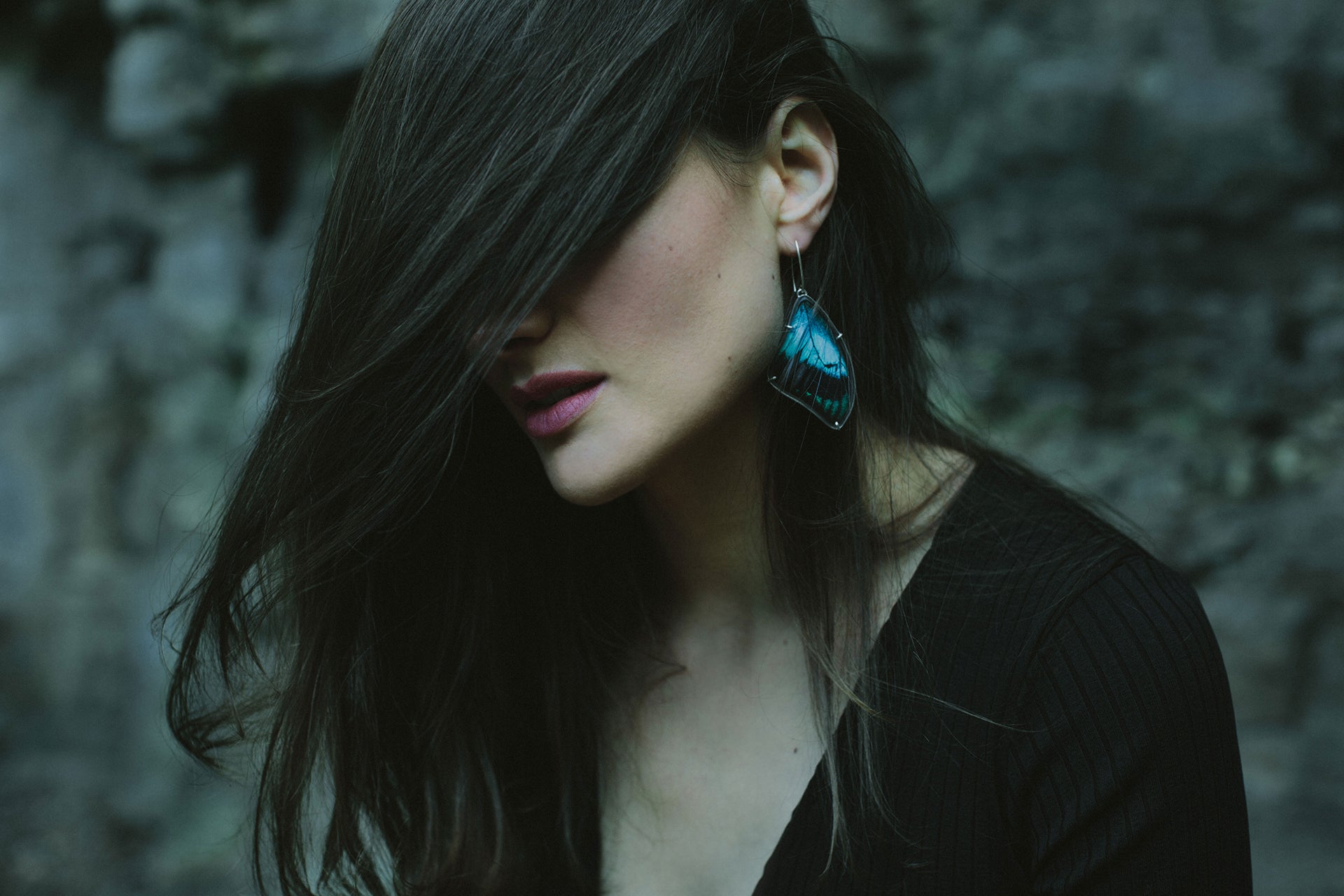 A woman with her dark hair swept over her face wears an aqua blue real butterfly wing earring. The image is dark and moody and set in front of a natural rock wall.