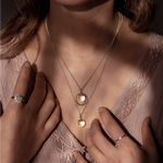 The Divination Necklace, which featuring a glowing quartz sphere on each pendant, are worn by a person wearing pink with dappled light landing on them.