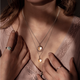 The Divination Necklace, which featuring a glowing quartz sphere on each pendant, are worn by a person wearing pink with dappled light landing on them.