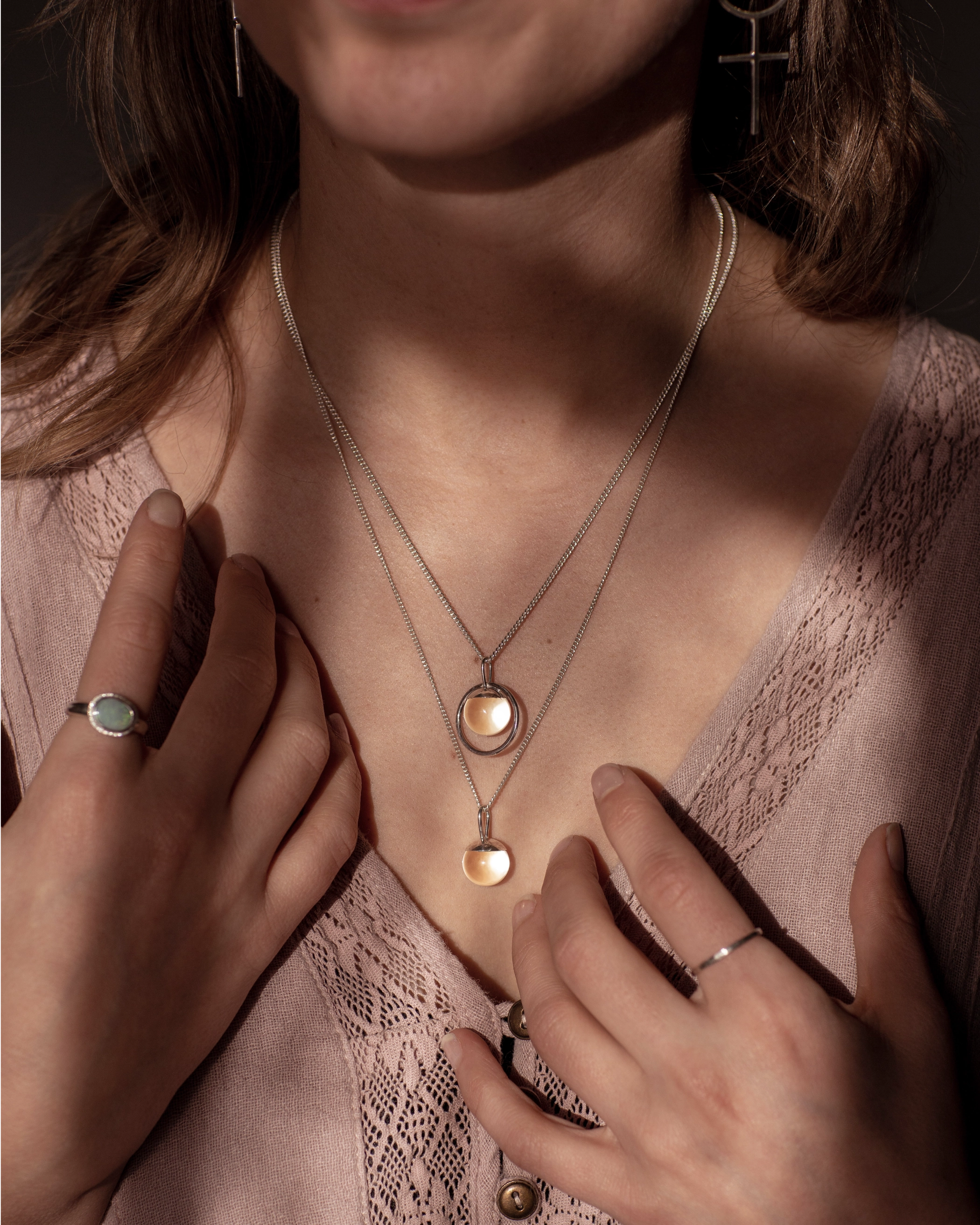 The Divination Necklace, which featuring a glowing quartz sphere on each pendant, are worn by a person wearing pink with dappled light landing on them.
