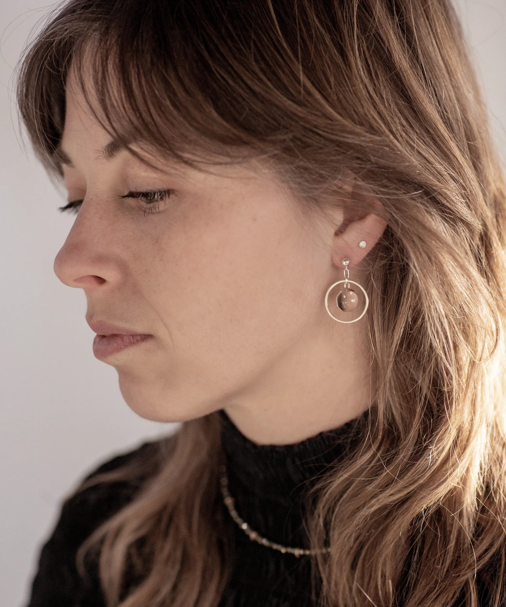 A person with a sweeping wolf-cut hairdo looking to the left wearing the Divination Stud Earrings, handmade by Emily Eliza Arlotte in Hobart, Tasmania. The earrings featuring a clear quartz crystal ball inside a hoop and hung from a stud fitting.
