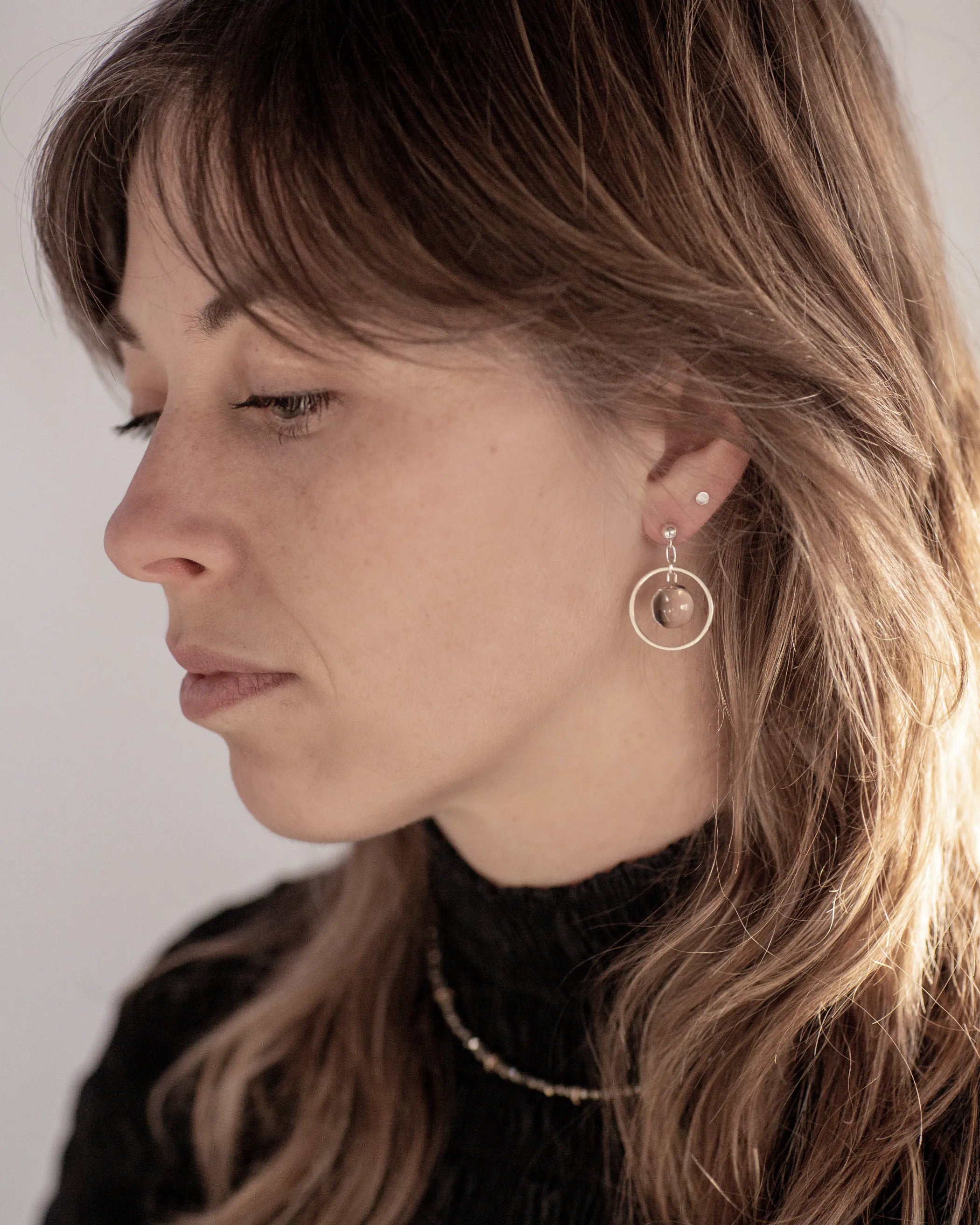 A person with a sweeping wolf-cut hairdo looking to the left wearing the Divination Stud Earrings, handmade by Emily Eliza Arlotte in Hobart, Tasmania. The earrings featuring a clear quartz crystal ball inside a hoop and hung from a stud fitting.