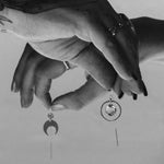 A black and white Close-up image of tattooed hands holding the Divination Thread earrings and the Lunar Thread Earrings with a plain background