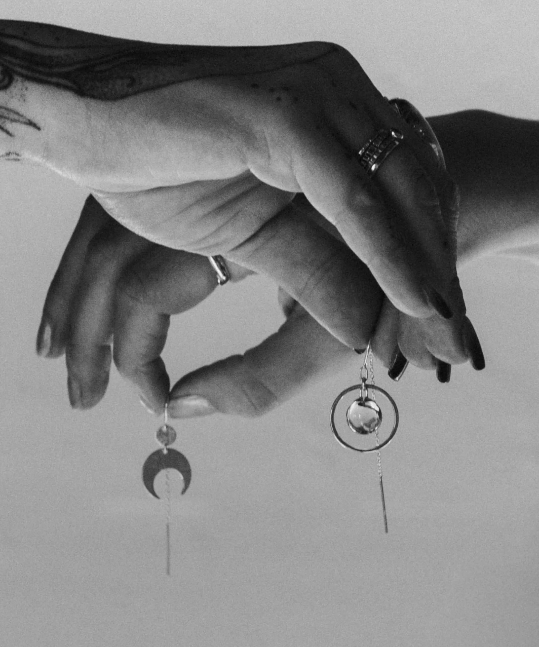 A black and white Close-up image of tattooed hands holding the Divination Thread earrings and the Lunar Thread Earrings with a plain background
