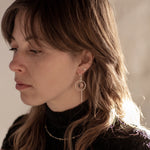 A person with brown wolf-cut hair wearing black high-neck textured top with the Divination Thread Earrings and the Pyrite Choker Necklace