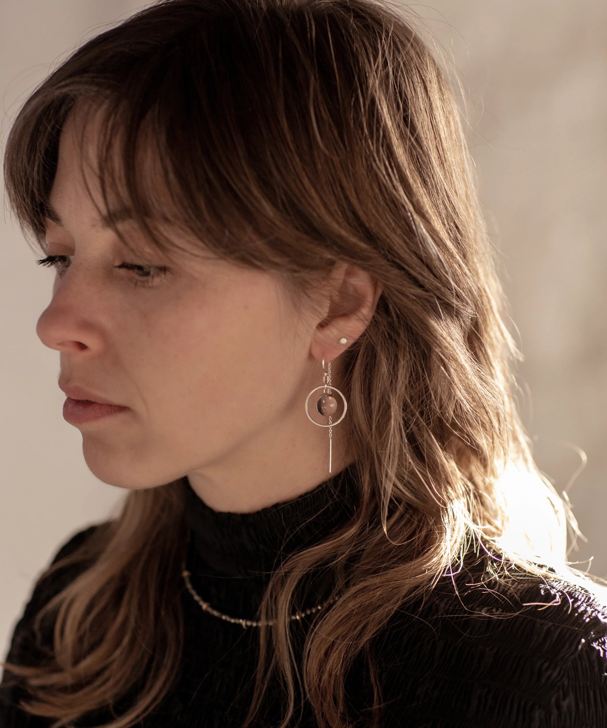 A person with brown wolf-cut hair wearing black high-neck textured top with the Divination Thread Earrings and the Pyrite Choker Necklace