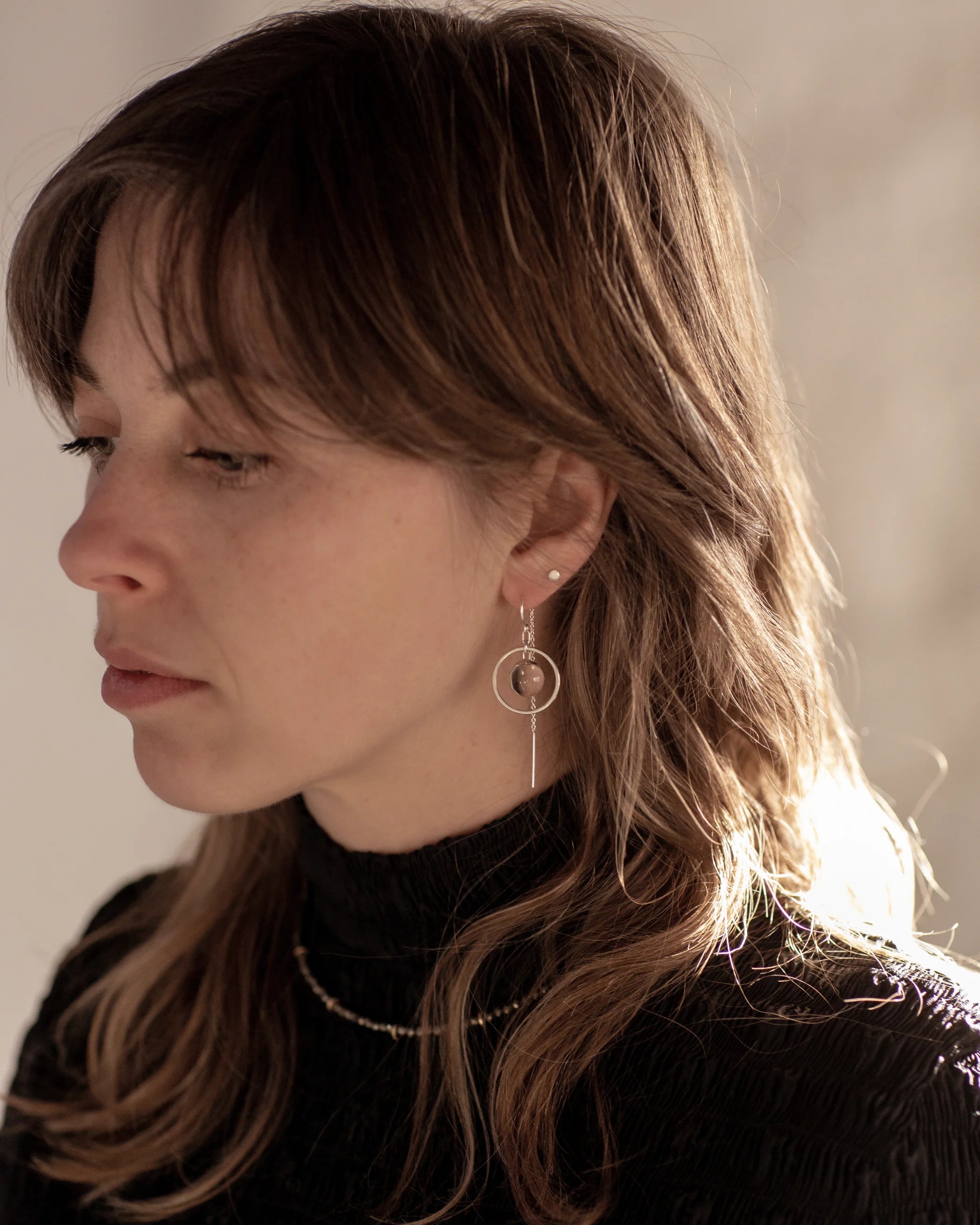 A person with brown wolf-cut hair wearing black high-neck textured top with the Divination Thread Earrings and the Pyrite Choker Necklace