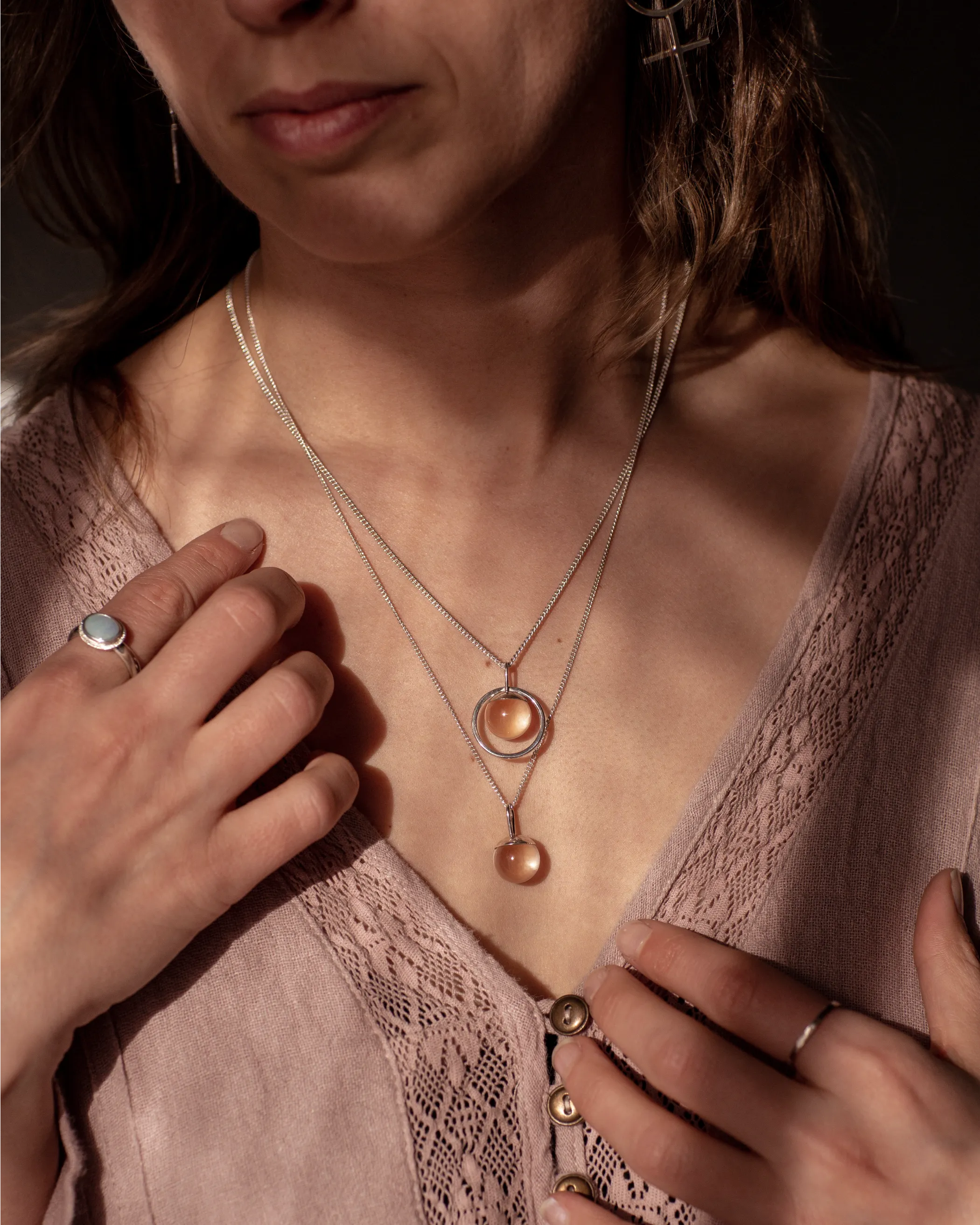 The Divination Necklaces, which feature a glowing quartz orb on each pendant, are worn by a person wearing pink with dappled light landing on them.