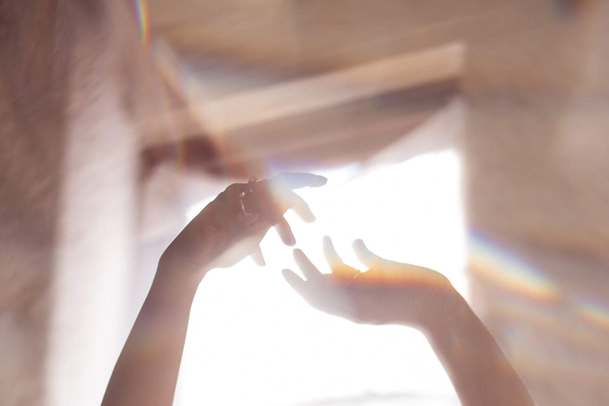 In an image made up of soft pinks and a gentle haze, two hands reach upwards in a relaxed, dance-like fashion in front of a window. 