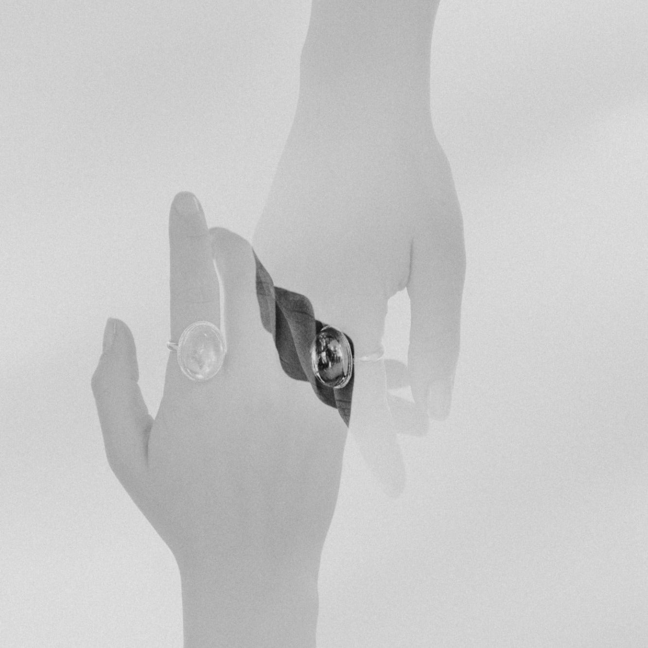Two hands, overlapping each other in a double exposure, wear a ring by Emily Eliza Arlotte. The image is soft and greyscale.