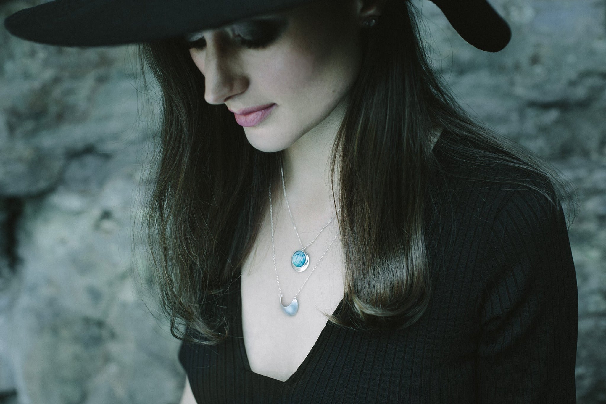 A woman wearing a large black hat looks downward from the camera. She is wearing two necklaces, a bright blue real butterfly wing pendant, and  luna moon necklace.