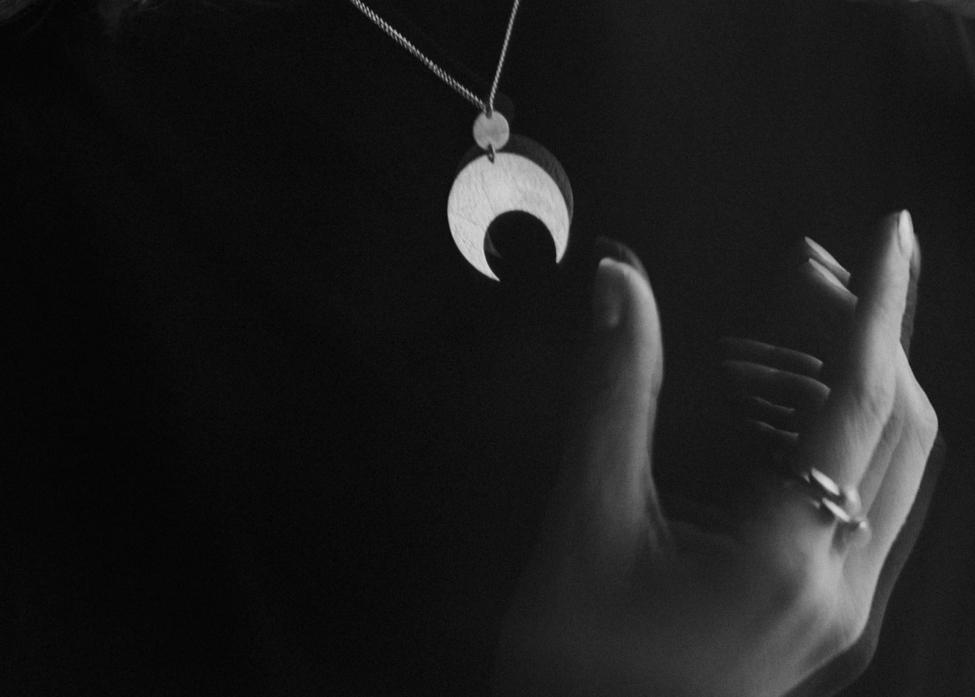 Hand holding a necklace with a crescent moon pendant by Emily Eliza Arlotte against a dark background. The image is double exposed.