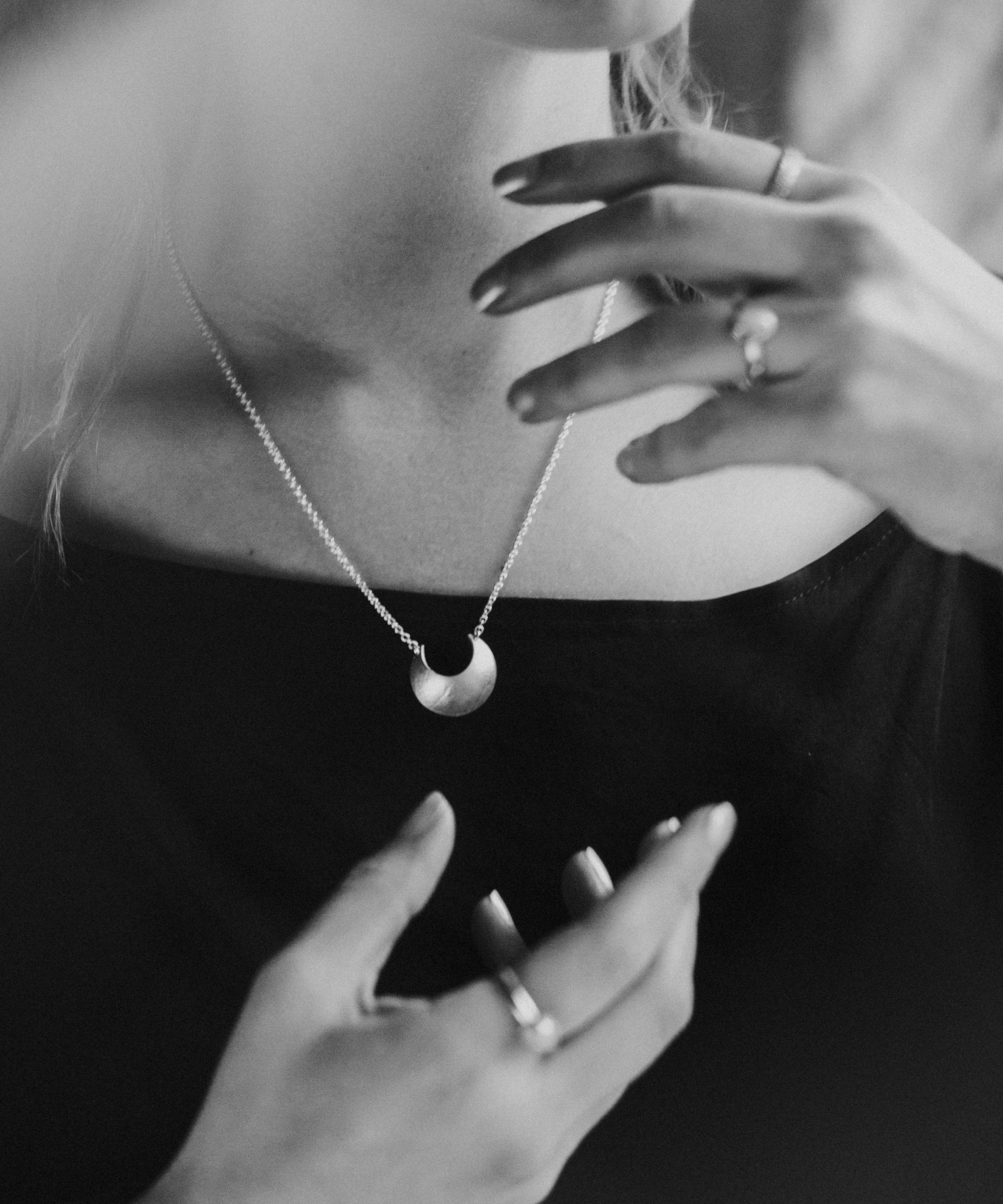 Black and white close-up of a person wearing  the Sterling Silver Luna Crescens necklace featuring a crescent moon by Emily Eliza Arotte. The persons hands are in a magical pose around the necklace.