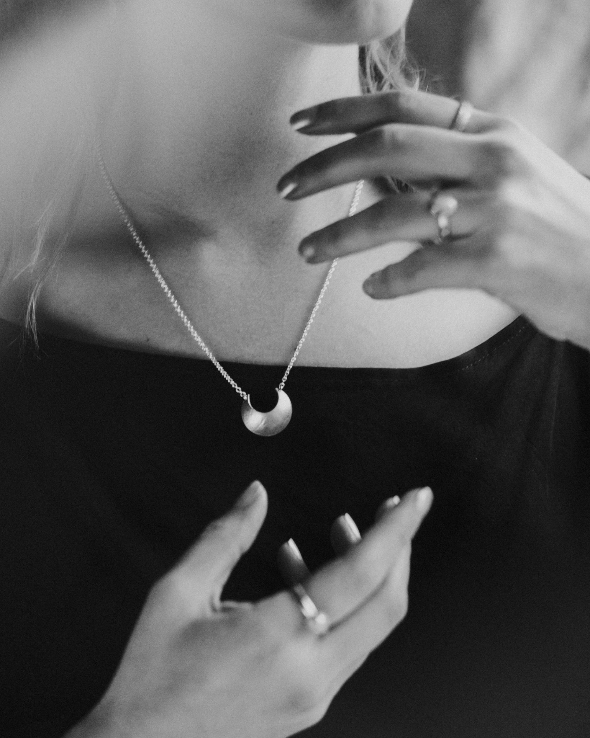 Black and white close-up of a person wearing  the Sterling Silver Luna Crescens necklace featuring a crescent moon by Emily Eliza Arotte. The persons hands are in a magical pose around the necklace.