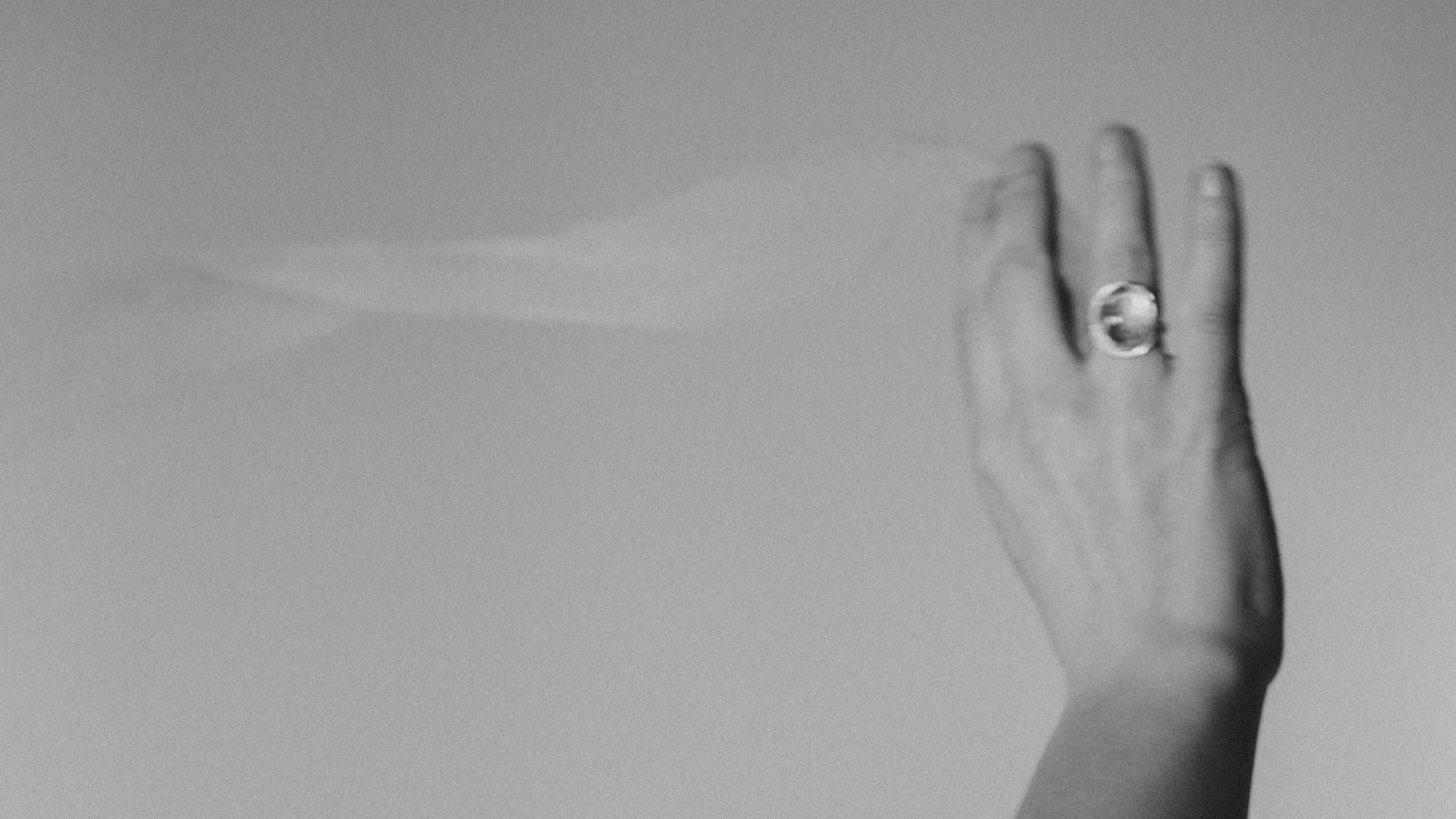 A hand wistfully waves a bridal veil through the air, while wearing an Emily Eliza Arlotte handmade bespoke engagement ring. The image is in soft, dreamy grey tones.