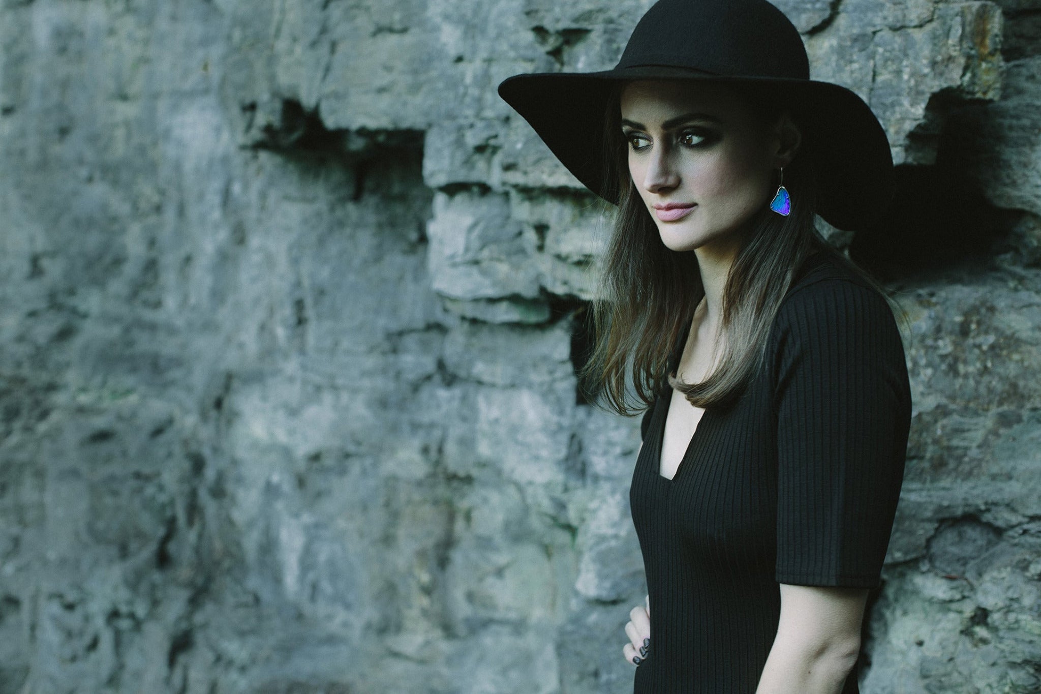 A woman wearing a large black hat stands in front of a rock face on Kunanyi / Mt Wellington. She is wearing a pair of bright blue real butterfly wing earrings.