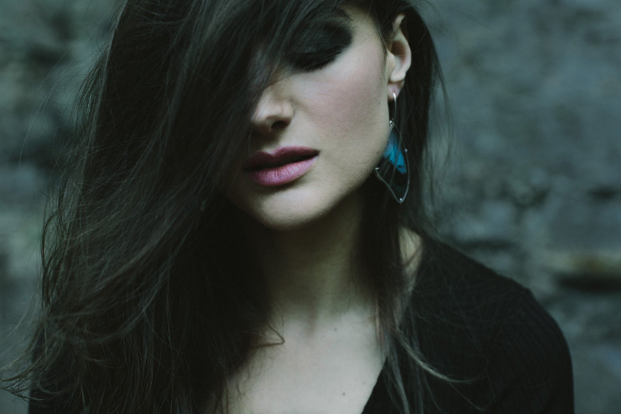 A woman with her dark hair dramatically swept across her face is wearing a pair of handmade real blue butterfly wing earrings.