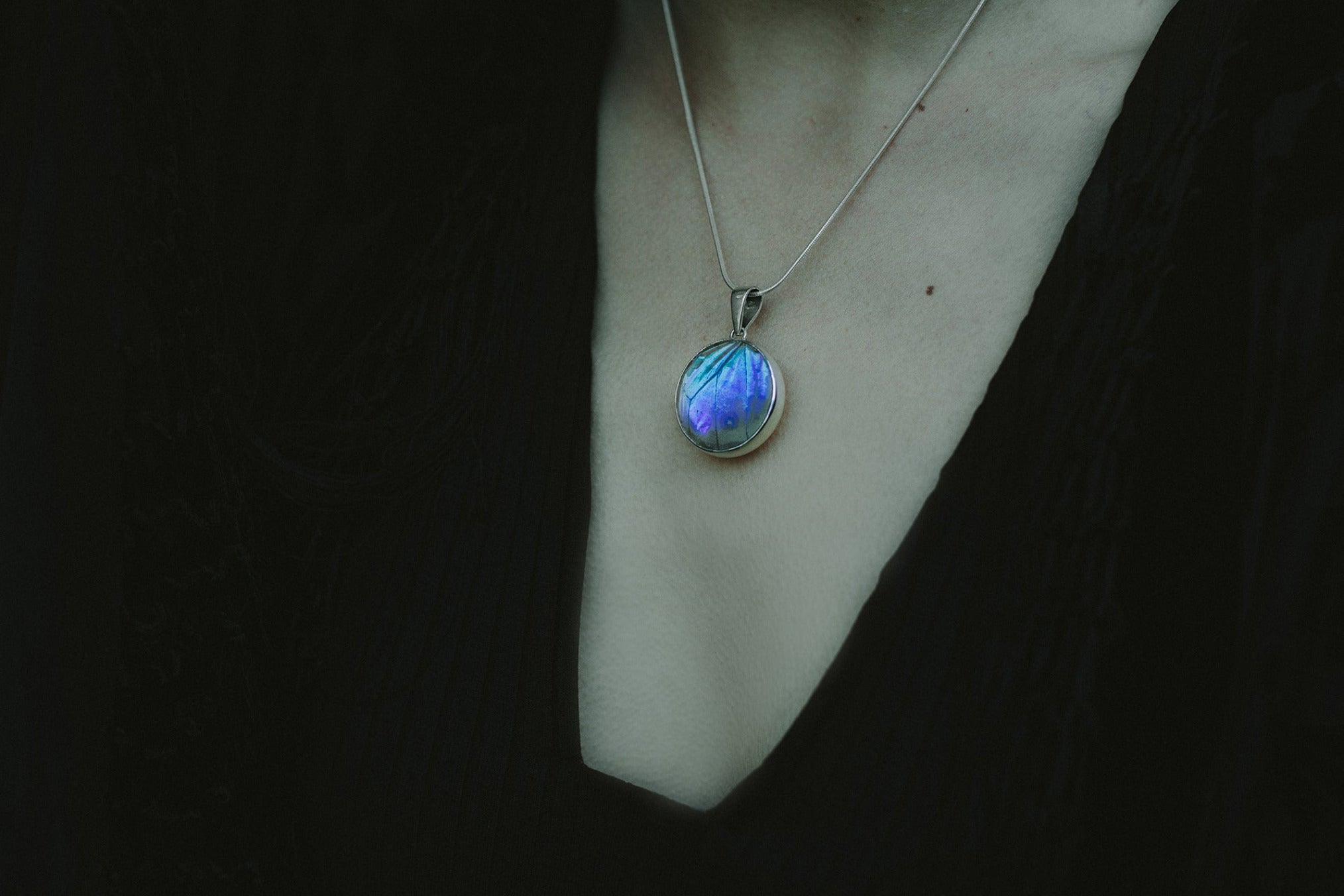 An image of a woman, only her chest is seen, wearing a handmade iridescent blue butterfly wing pendant set in sterling silver and glass.