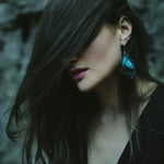 A woman has her dark hair dramatically swept across her face, showing a large blue real butterfly wing earring.