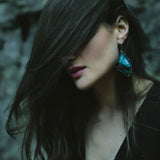 A woman has her dark hair dramatically swept across her face, showing a large blue real butterfly wing earring.