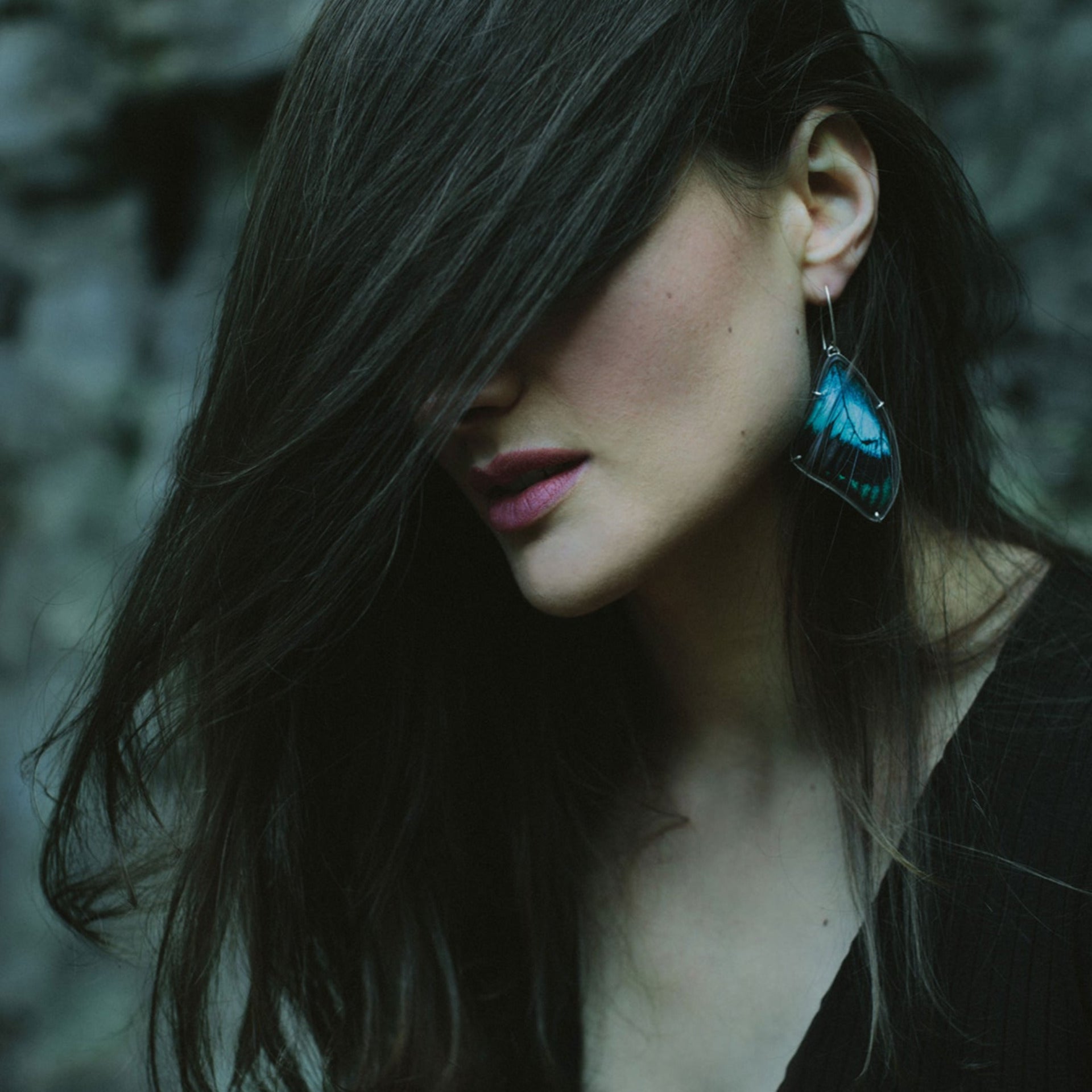 A woman has her dark hair dramatically swept across her face, showing a large blue real butterfly wing earring.