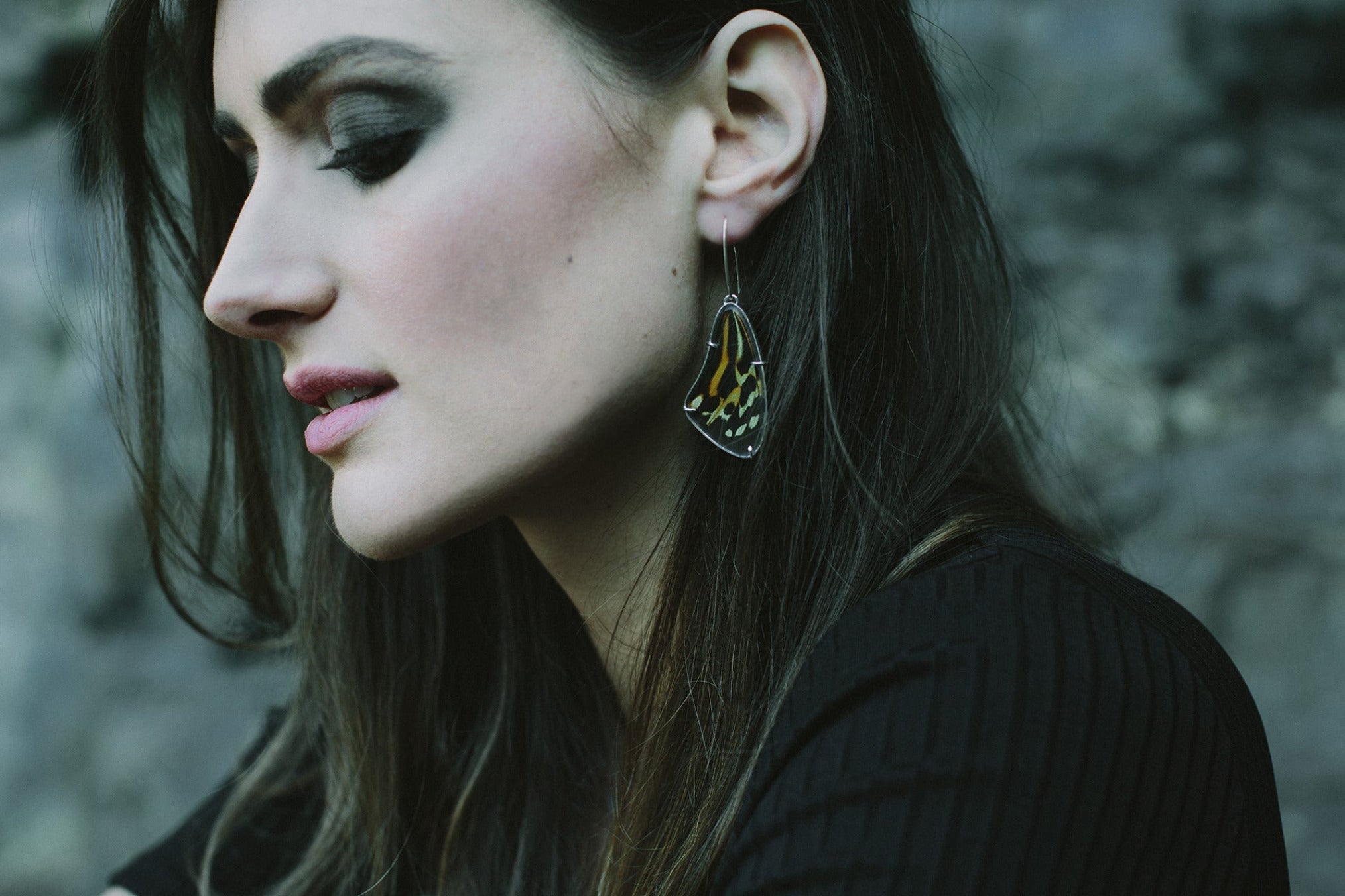 Laska wears  pair of handmade real buuterfly wing earrings. The earrings are orange and black striped and hang low from the ear.