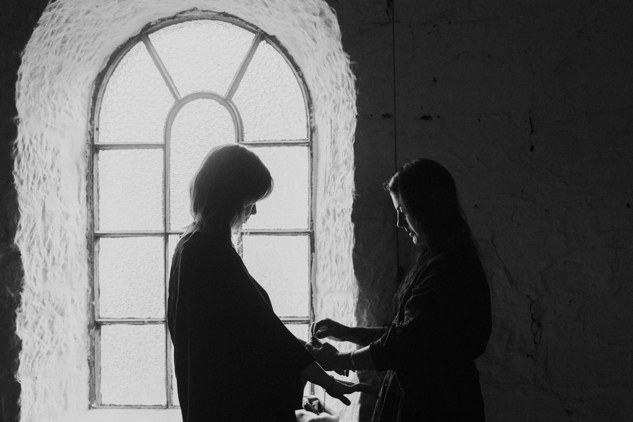 Emily Eliza Arlotte stands in front of an arch window, with a textured wall. They are placing jewellery on the model, Eden.