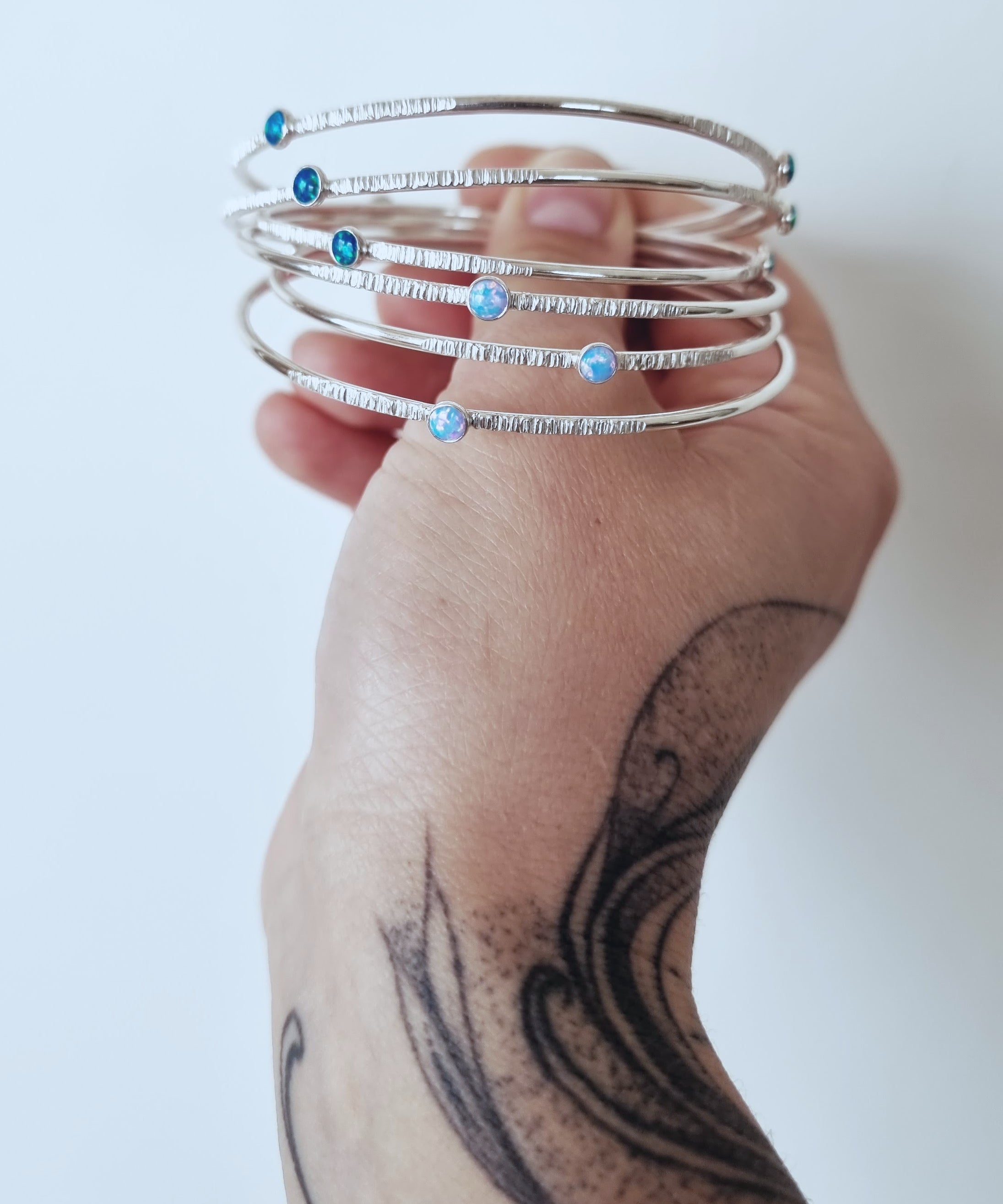 A tattooed hand holds a collection of Sterling Silver Opal Bangles by Emily Eliza Arlotte