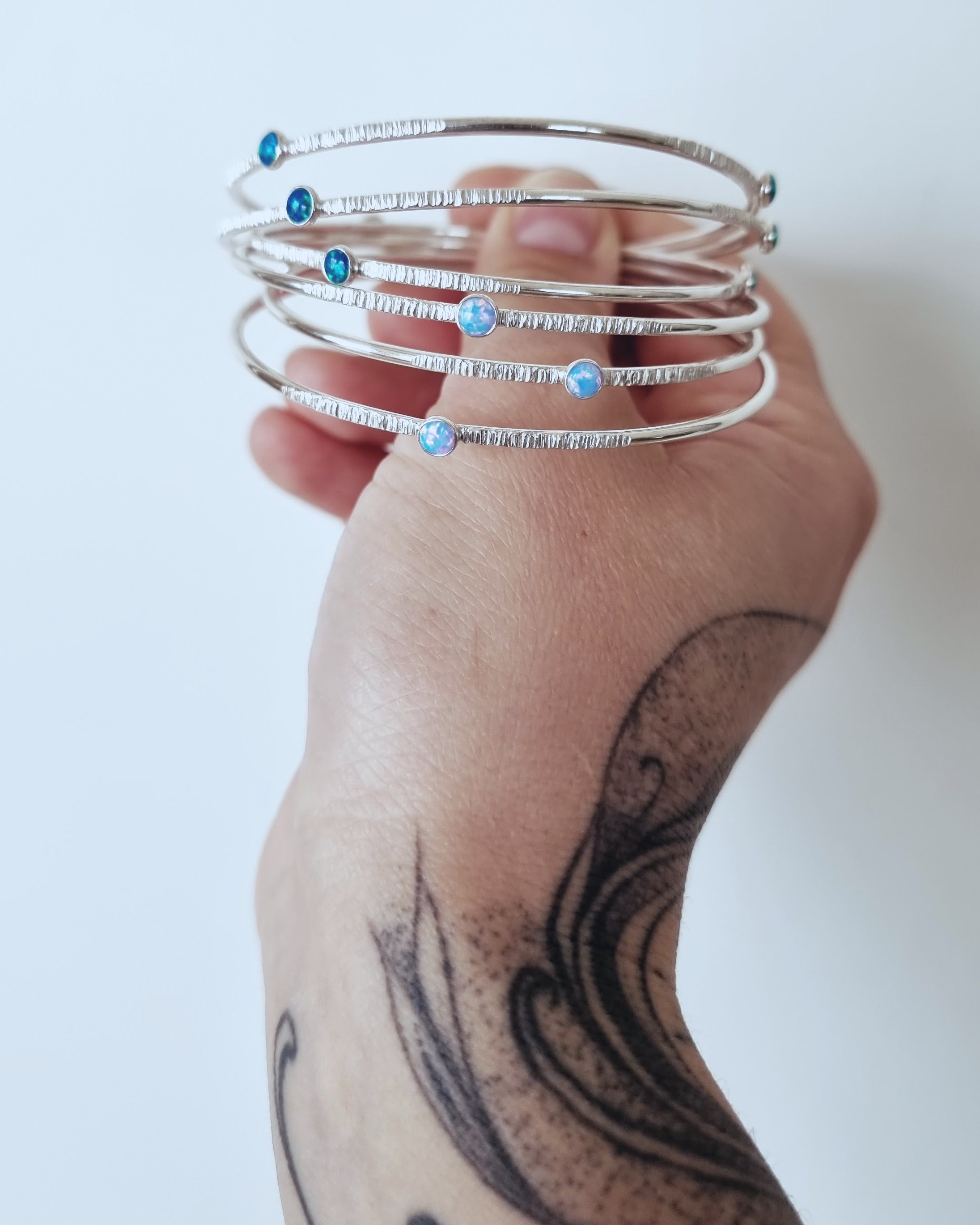 A tattooed hand holds a collection of Sterling Silver Opal Bangles by Emily Eliza Arlotte